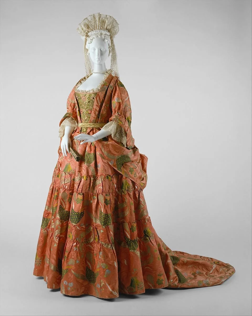 A mannequin dressed in elaborate 18th-century style gown with a long train, gold and pink fabric, lace accents, and a matching headdress.