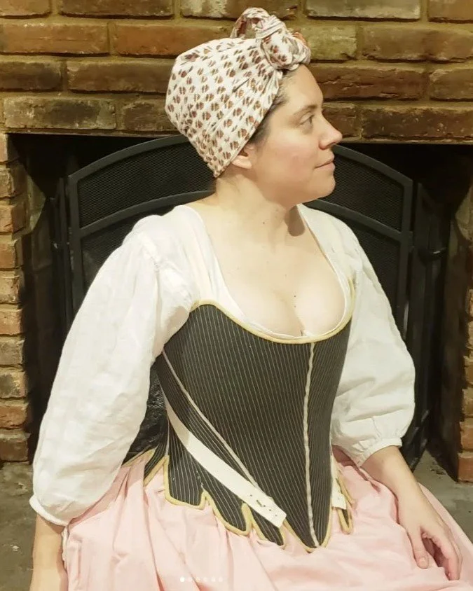 Sarah is sitting in front of a fireplace wearing her newly completed 1760s stays, made up in dark green fabric with cream binding and straps. This is a clear view of the front of her stays, made up to the custom arc-method pattern by Rebecca.