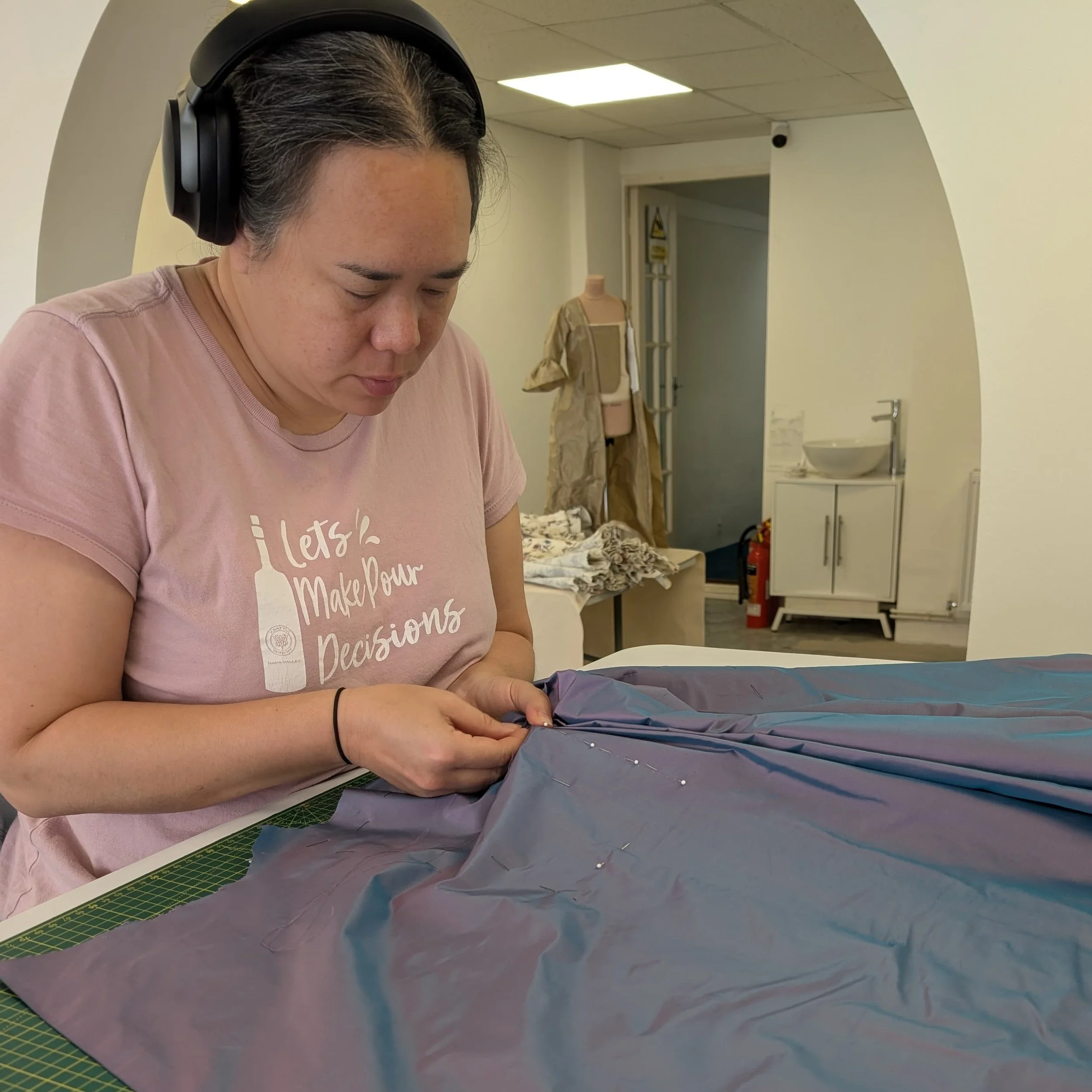 A person wearing a black helmet is working with fabric or material at a table, with sewing pins and cutting mats visible in an indoor setting.