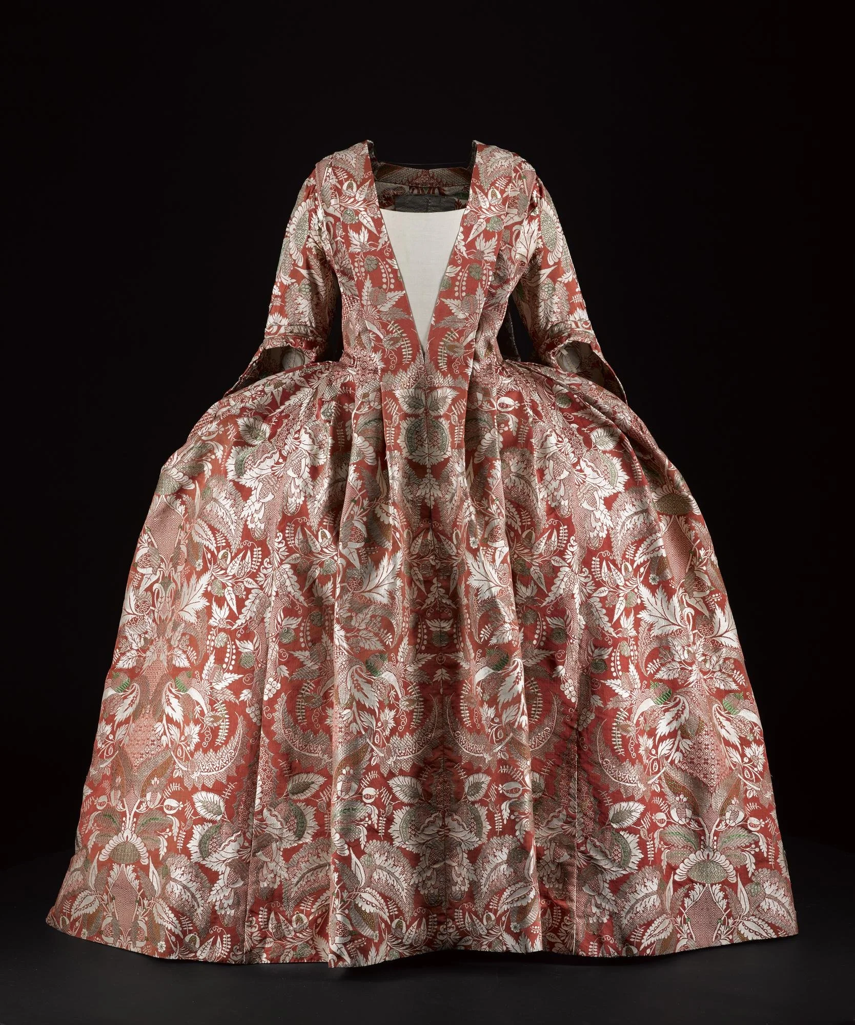 A vintage dress with intricate floral and leaf patterns in red, white, silver, and green hues, displayed against a black background.