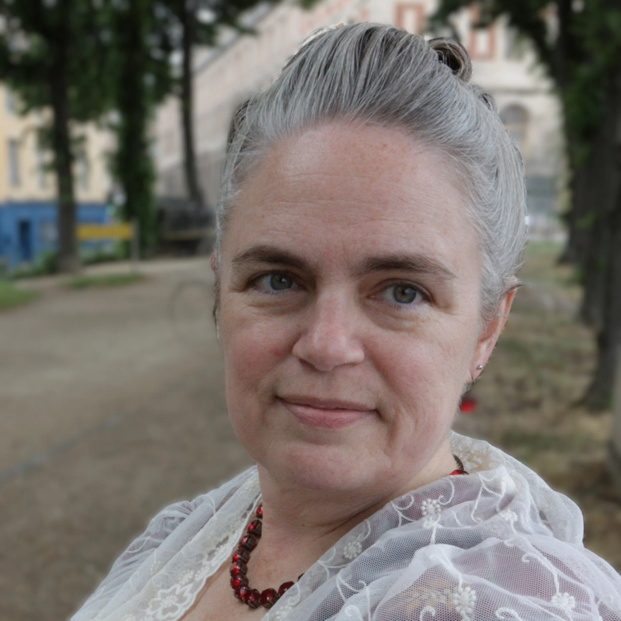 Close-up of Rebecca with powdered and pomaded hair in a 1760s style, wearing a red parure set of jewellery and a 1760s lace shawl, outdoors with blurred trees and buildings in the background.