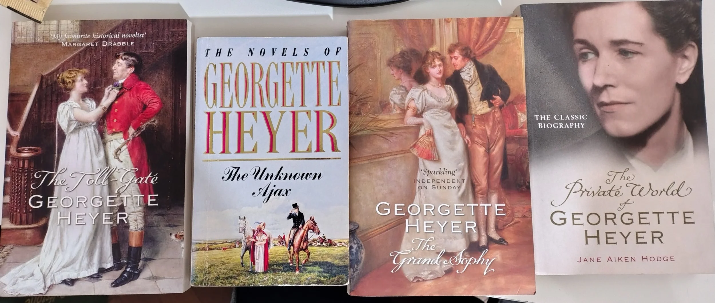 Four books by Georgette Heyer. The first book has a cover illustration of a woman in a white dress talking to a man in a red coat at the bottom of a staircase. The second book's cover features a painting of three women and a man in historical attire, with a background of potted plants. The third book shows a cover illustration of a man in formal historical attire talking to two women in a room with curtains. The fourth book's cover has a close-up portrait of a woman with short dark hair, titled "The Private World of Georgette Heyer."