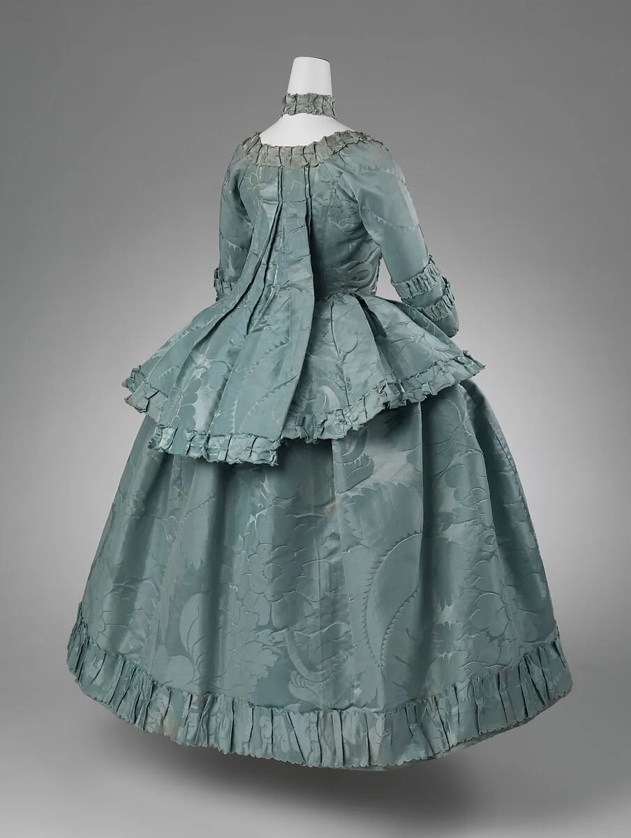 A vintage teal-colored women's gown with a patterned brocade fabric, a fitted bodice, voluminous skirt, ruffled trim at the hem and sleeves, and a matching peplum, displayed on a mannequin against a plain background.