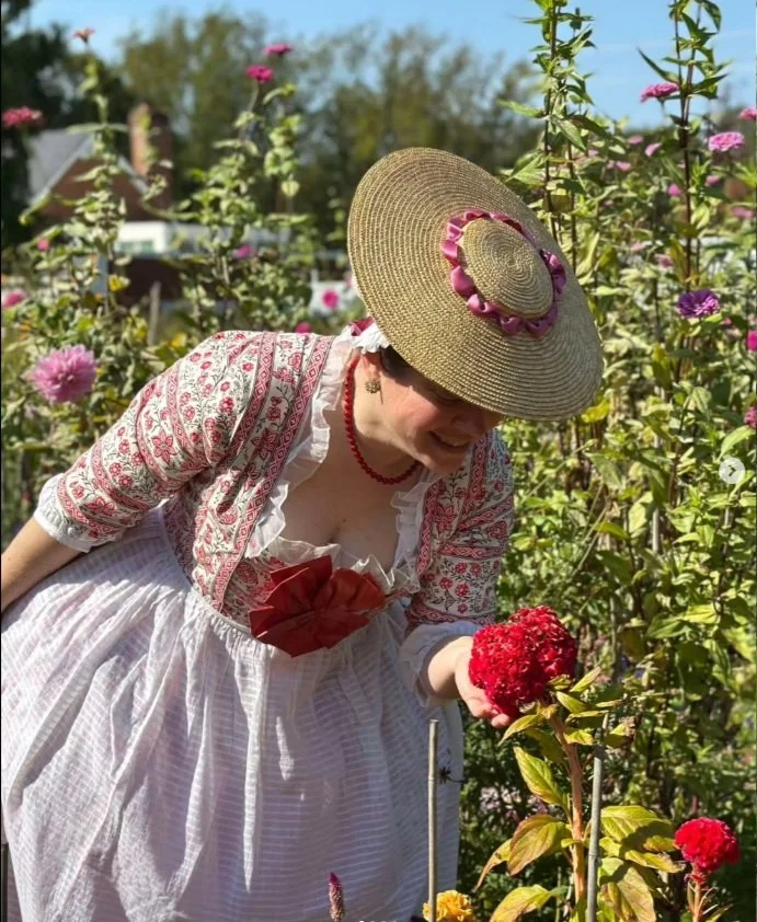 Sarah wearing her new floral-printed cotton longsack gown, perfectly fitted over her new 1760s stays made according to the custom arc-method pattern drafted by Rebecca. Sarah is in a sunny garden, bending to smell the fragrance of a large red flower.