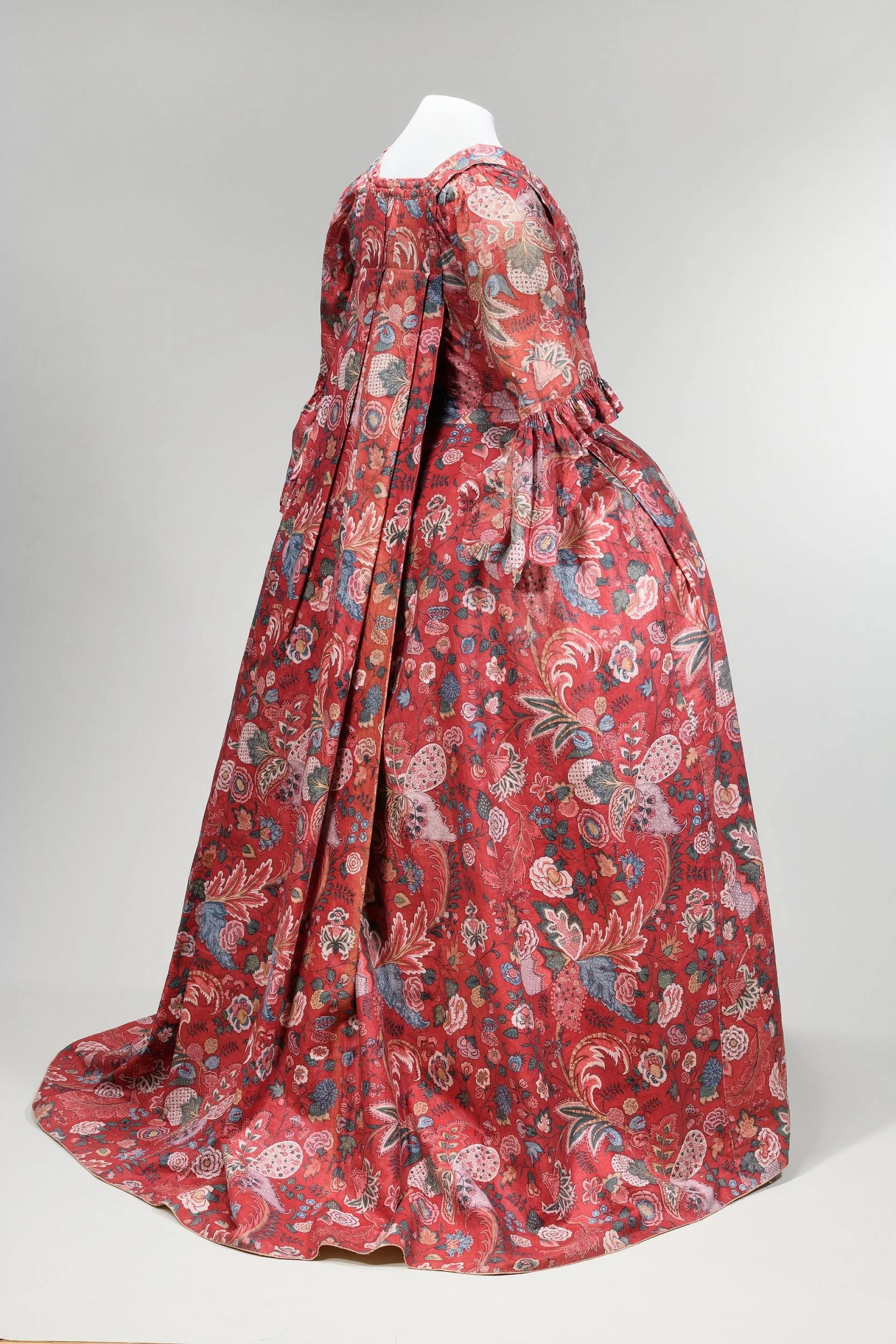 A mannequin dressed in a long, floral-patterned, red gown with puffed sleeves and a peplum waist, displayed against a plain light-colored background.