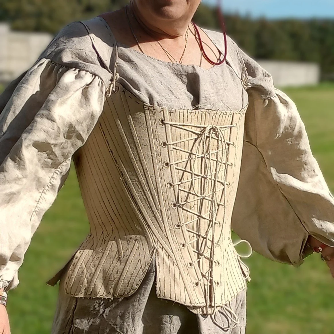 Tania stands outdoors wearing the first mock-up for her new 1740s stays, made from the custom arc-method pattern drafted by Rebecca. This is the front view. She is also wearing a natural-coloured linen shift with very full sleeves.