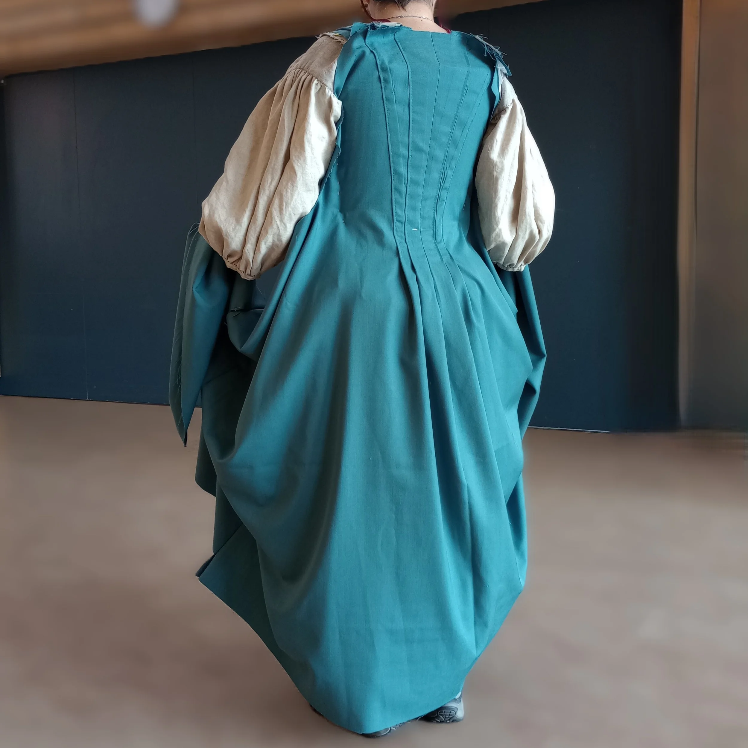 Back view of a person in historical costume, wearing a teal blue dress with a fitted bodice and full skirt, and beige puffed sleeves, standing in front of a dark background.