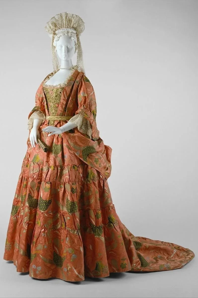 A mannequin dressed in a luxurious, vintage-style, peach and gold patterned gown with a long train, intricate lace sleeves, and a pearl necklace, with an elaborate lace headdress.
