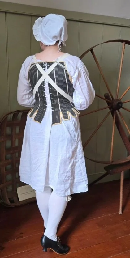 Sarah stands with her back to the viewer, showing the back of her newly completed dark green stays, made up to the custom arc-method pattern by Rebecca. She is also wearing a white linen shift, white linen cap, cream stockings and black shoes.