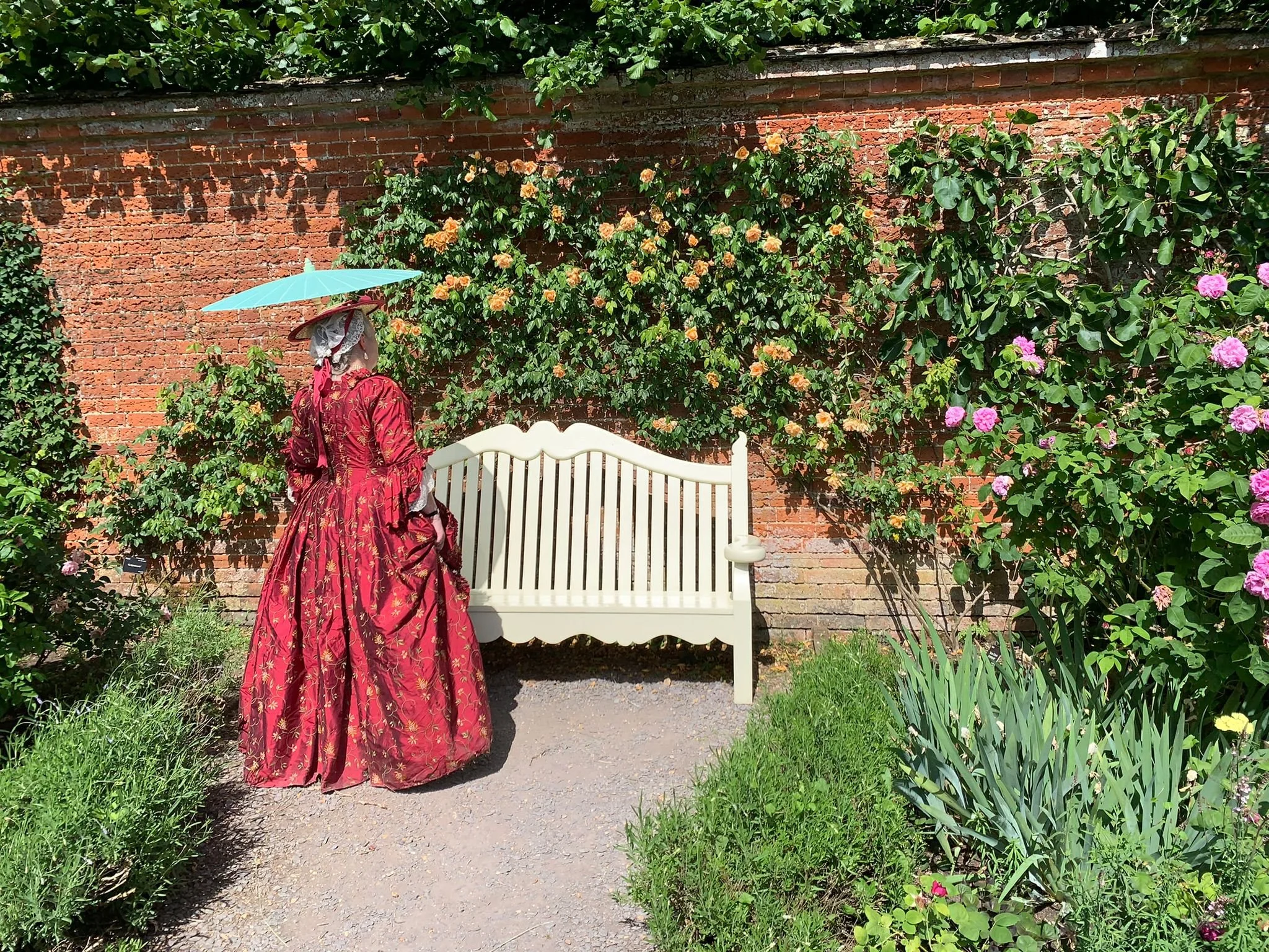 A woman in an embroidered red silk 18th century gown stands before a cream wooden bench with her back to us as she admires climbing roses on a red brick garden wall.