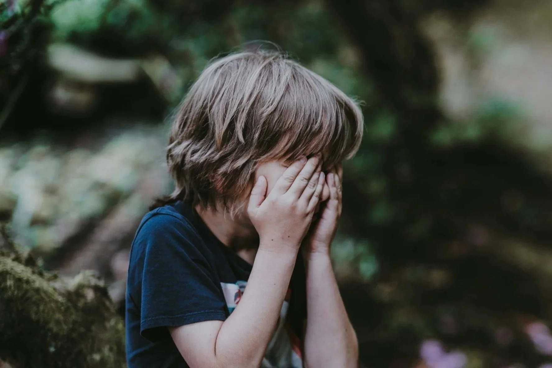 A young person with light brown hair, wearing a dark blue shirt, covering their face with their hands in a forest setting with green foliage in the background.