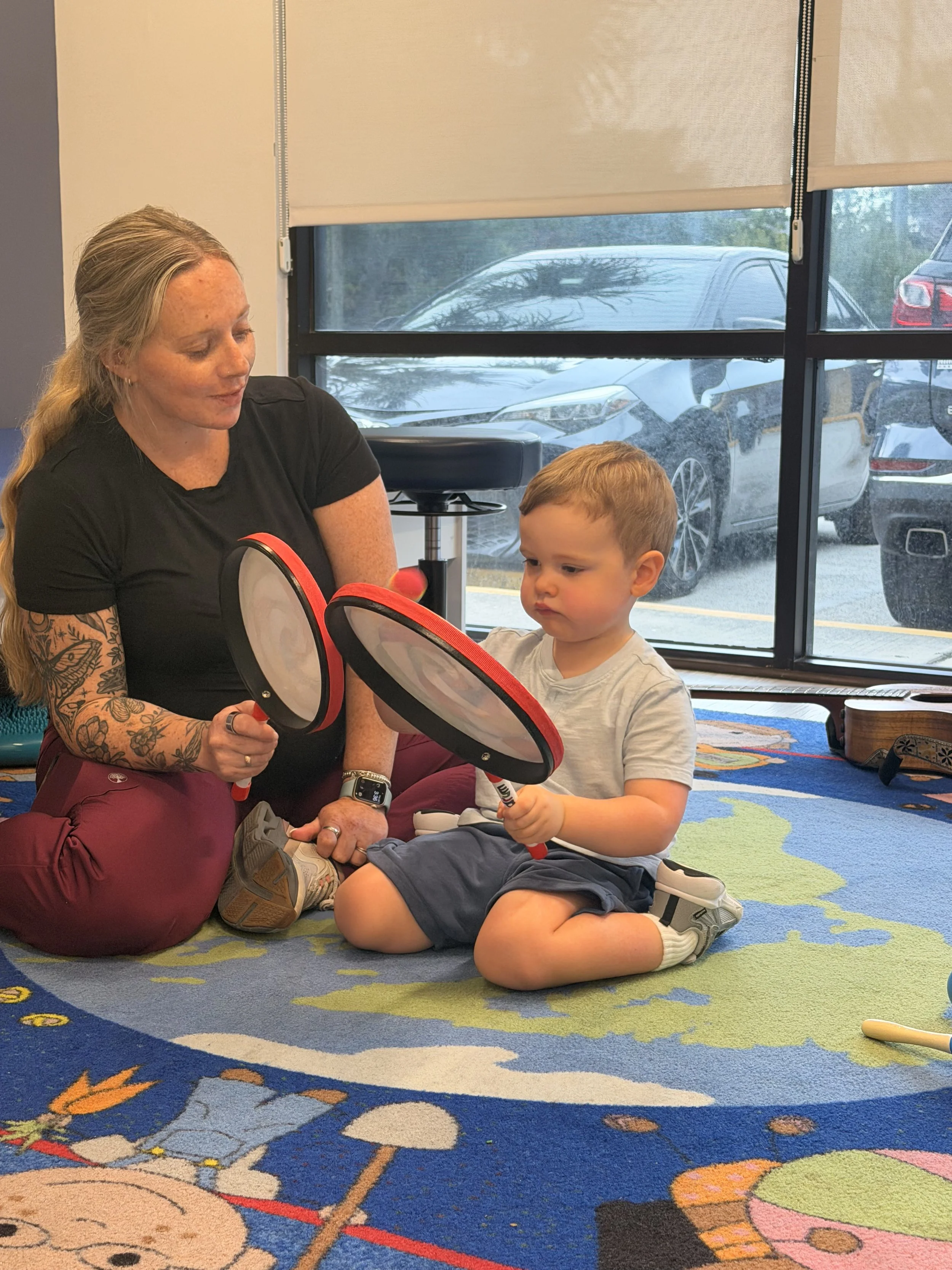 A woman with long blonde hair and tattoos on her arm is kneeling beside a young boy with short red hair, sitting on a colorful carpet, holding a toy and looking at it. They are indoors near a window with blinds, and parked cars are visible outside.