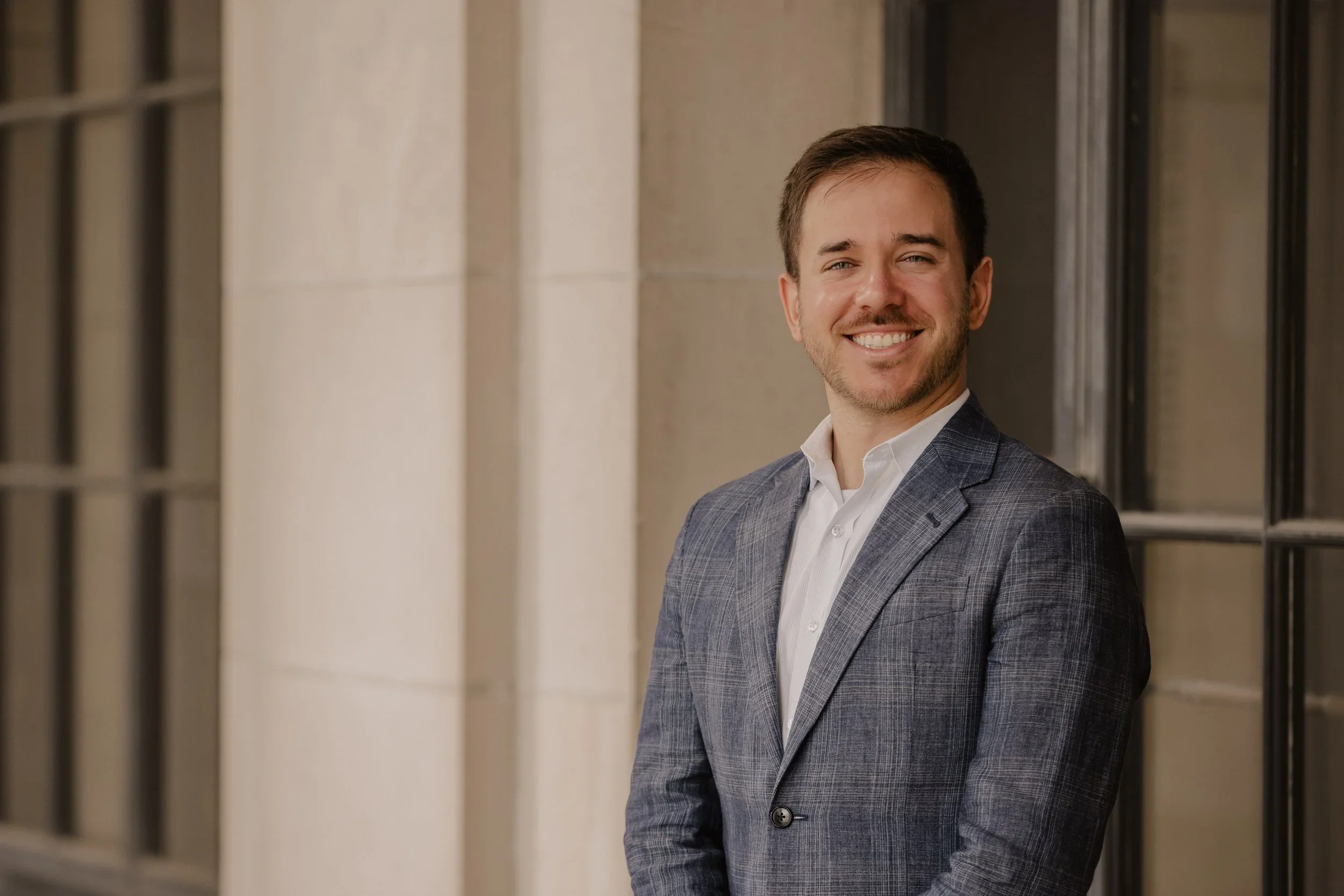 A smiling man in a gray checkered suit standing outside near a building with large windows.