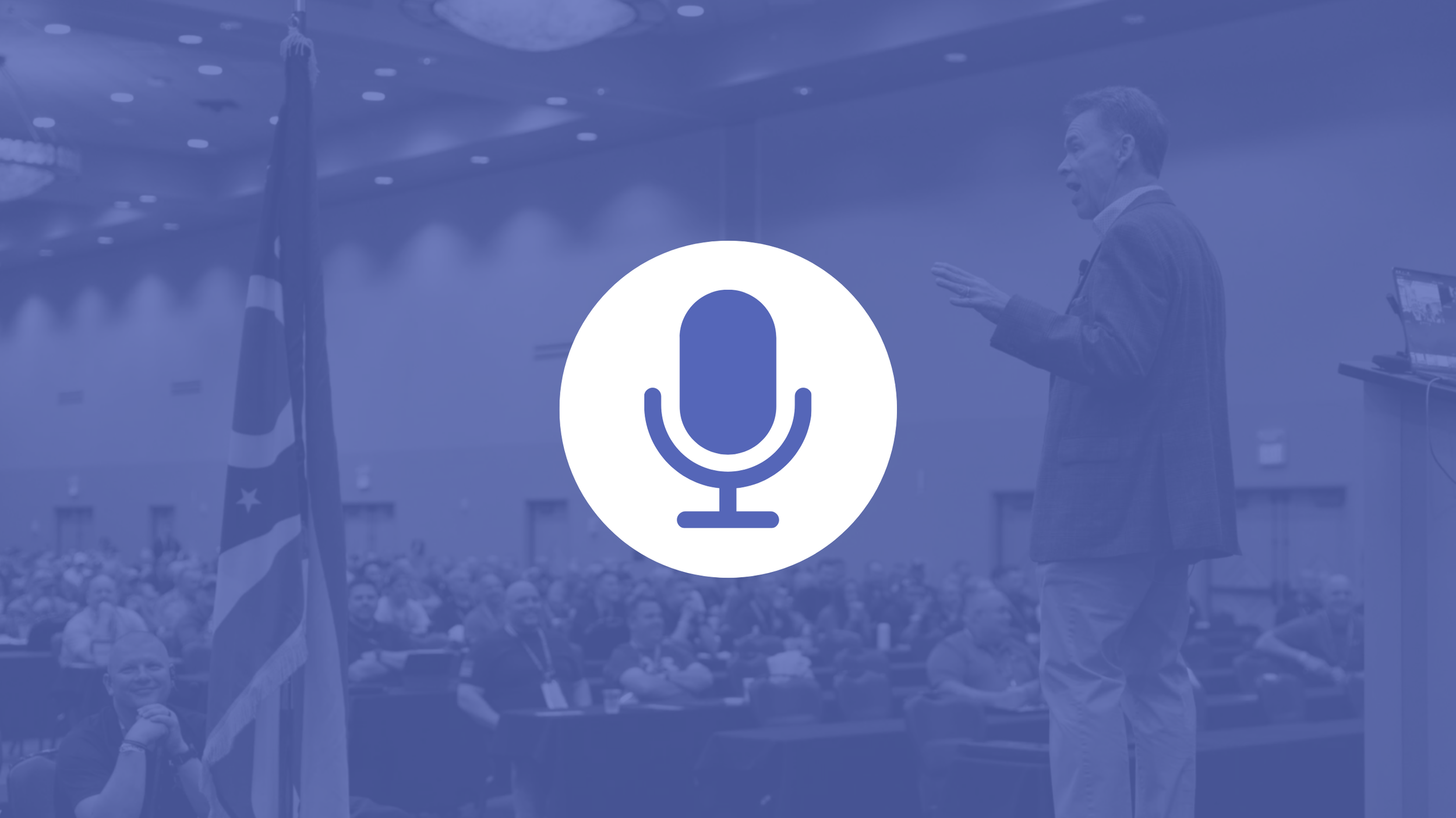 A speaker addressing a large audience in a conference hall with a British and American flag in the background, during a presentation or speech.