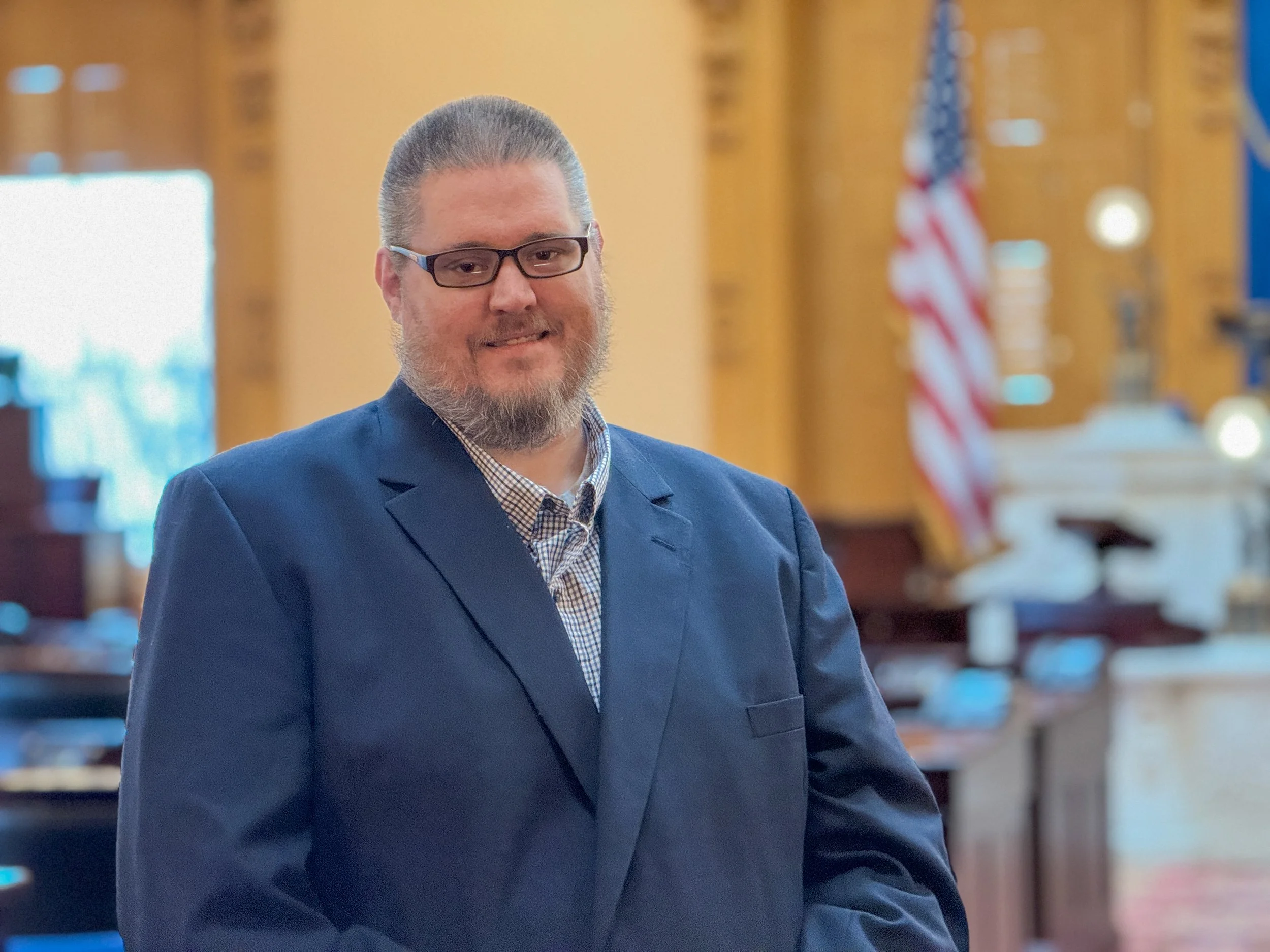 A man with glasses and a beard, wearing a navy suit and checkered shirt, standing in a room with wooden walls, American flag, and judge's bench in the background.