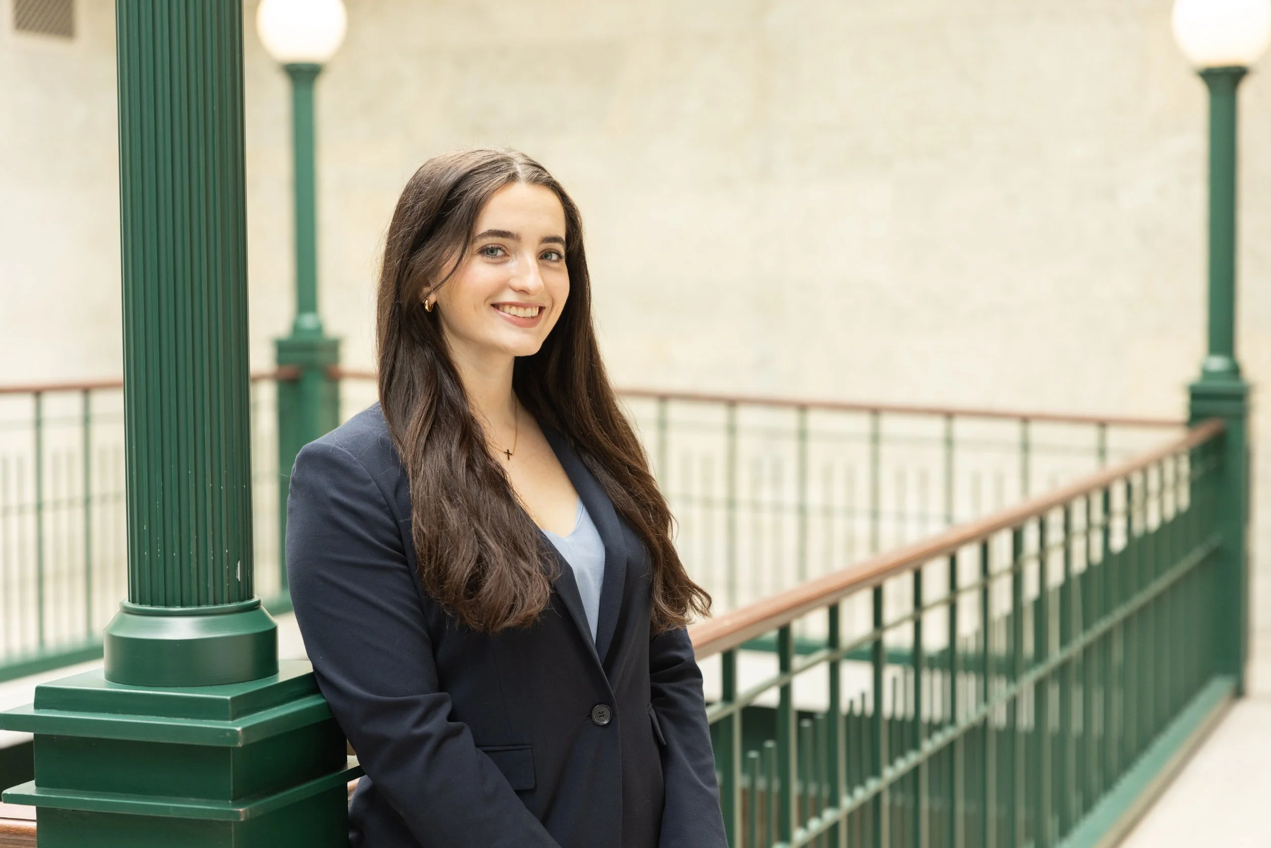 A young woman with long brown hair, wearing a navy blazer and light blue top, smiling and leaning against a green railing on a bridge or balcony.