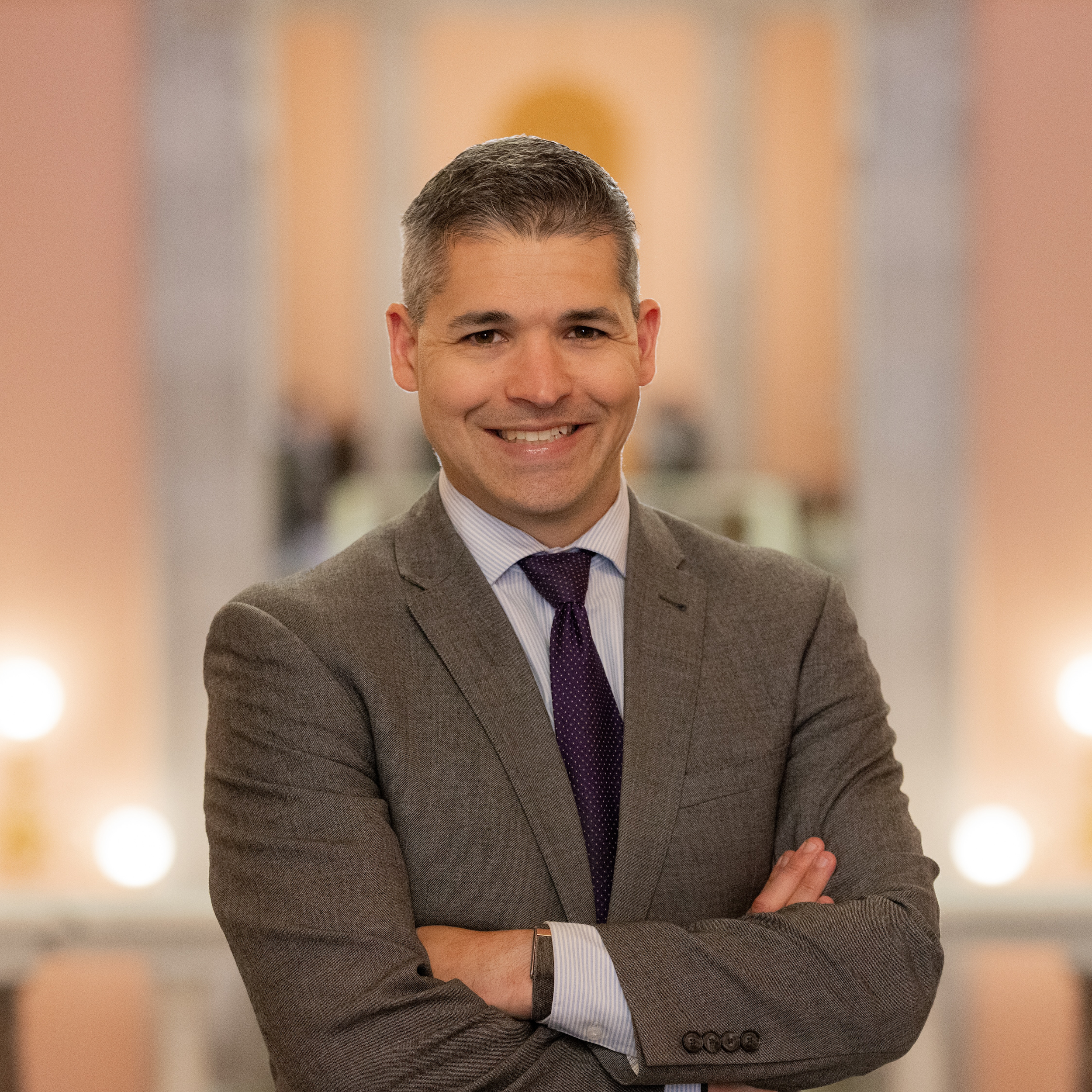 A smiling man in a gray suit with arms crossed, standing in a well-lit indoor setting with blurred warm-colored background lights.