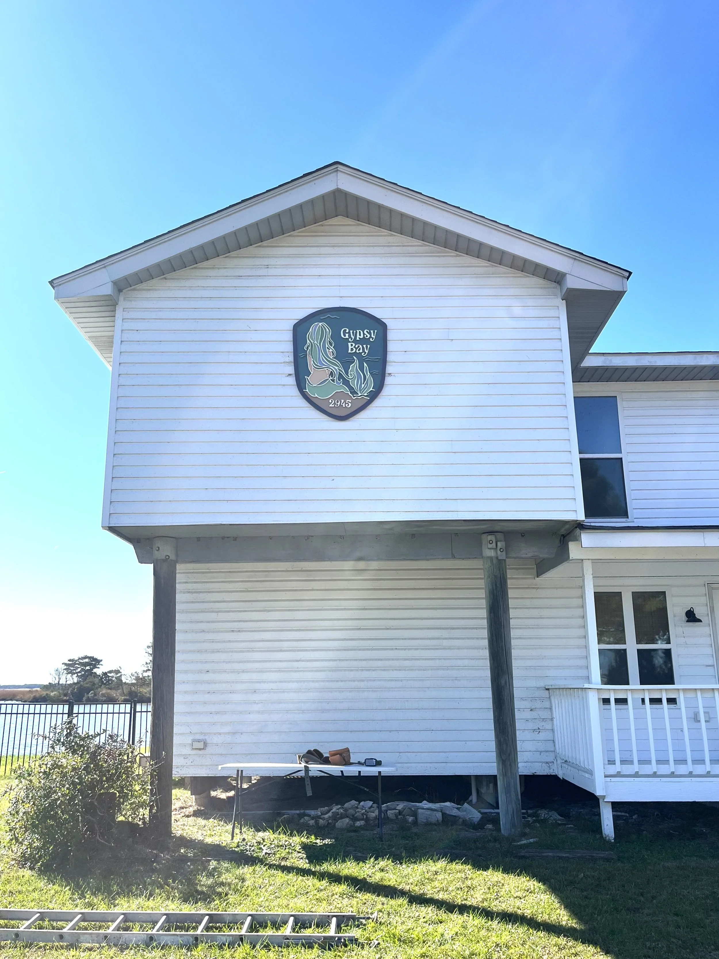 White house with a sign saying 'Gypsy Bay' and a mermaid illustration, situated in an outdoor setting with a grassy yard, some garden tools, and a fence in the background.