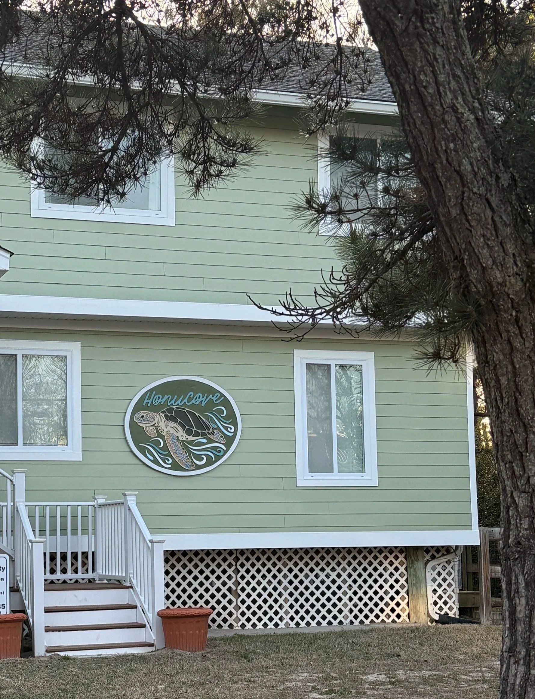 A light green house with white trim, a deck with stairs, and a large circular sign featuring a turtle and the word 'Honucape' on the front.