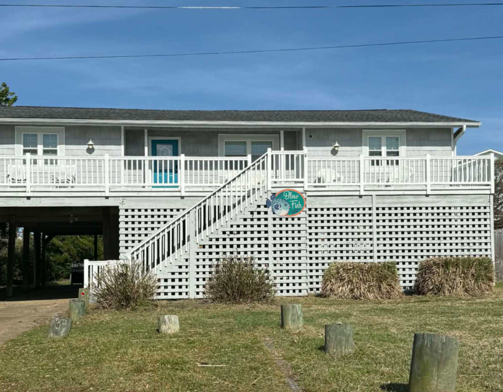 A two-story house with white wooden siding, a dark gray shingled roof, a front deck with white railing, and stairs leading to the yard below; a tree on the left side, a sign for Blow Fish, and a blue sky with a few clouds.