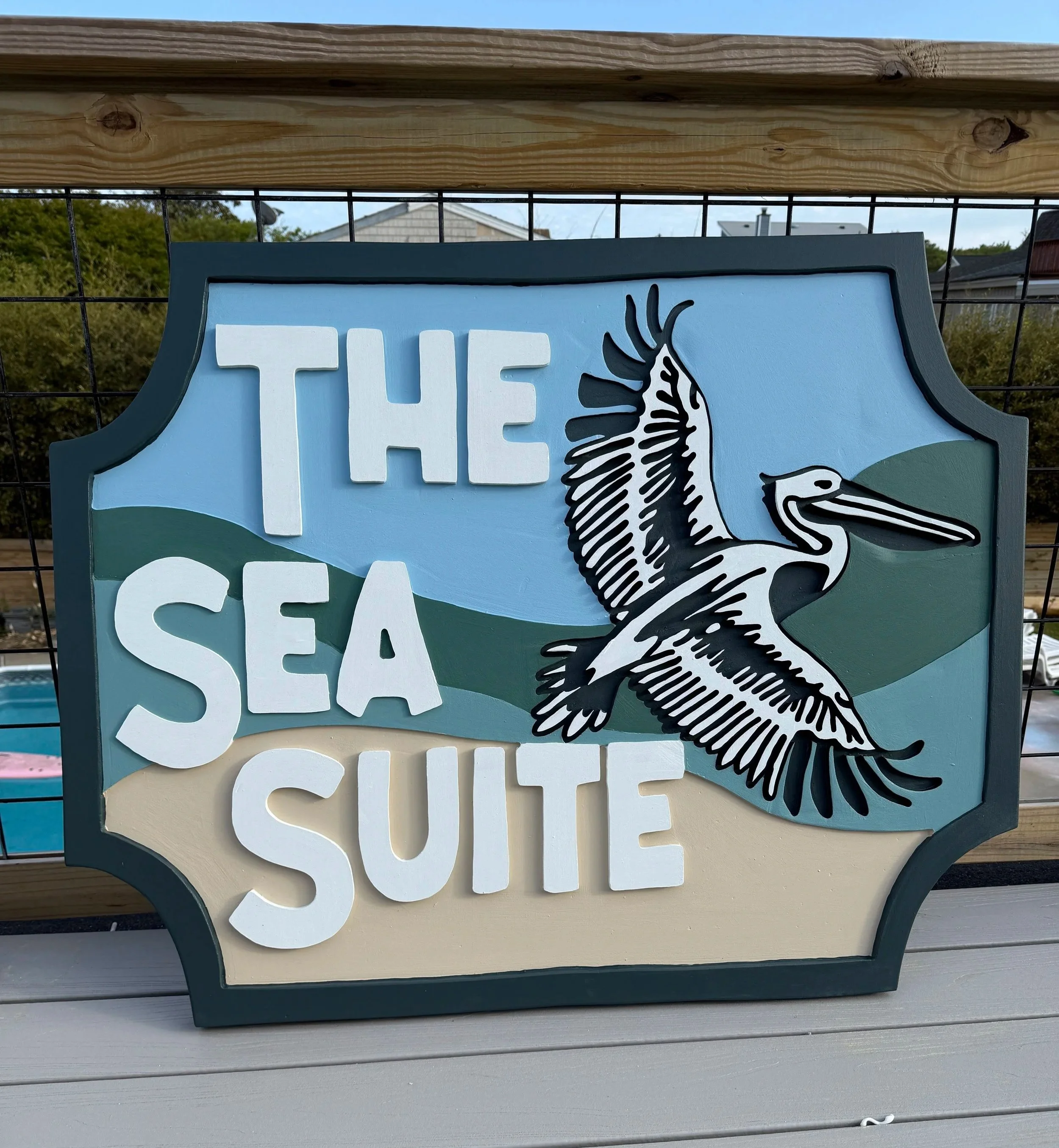 Close-up of a decorative sign with the partially visible words 'THE SEAFO'. The sign includes a black and white illustration of a seagull and is set against a backdrop of a wire fence, a wooden frame, and residential houses with trees and a cloudy sky.