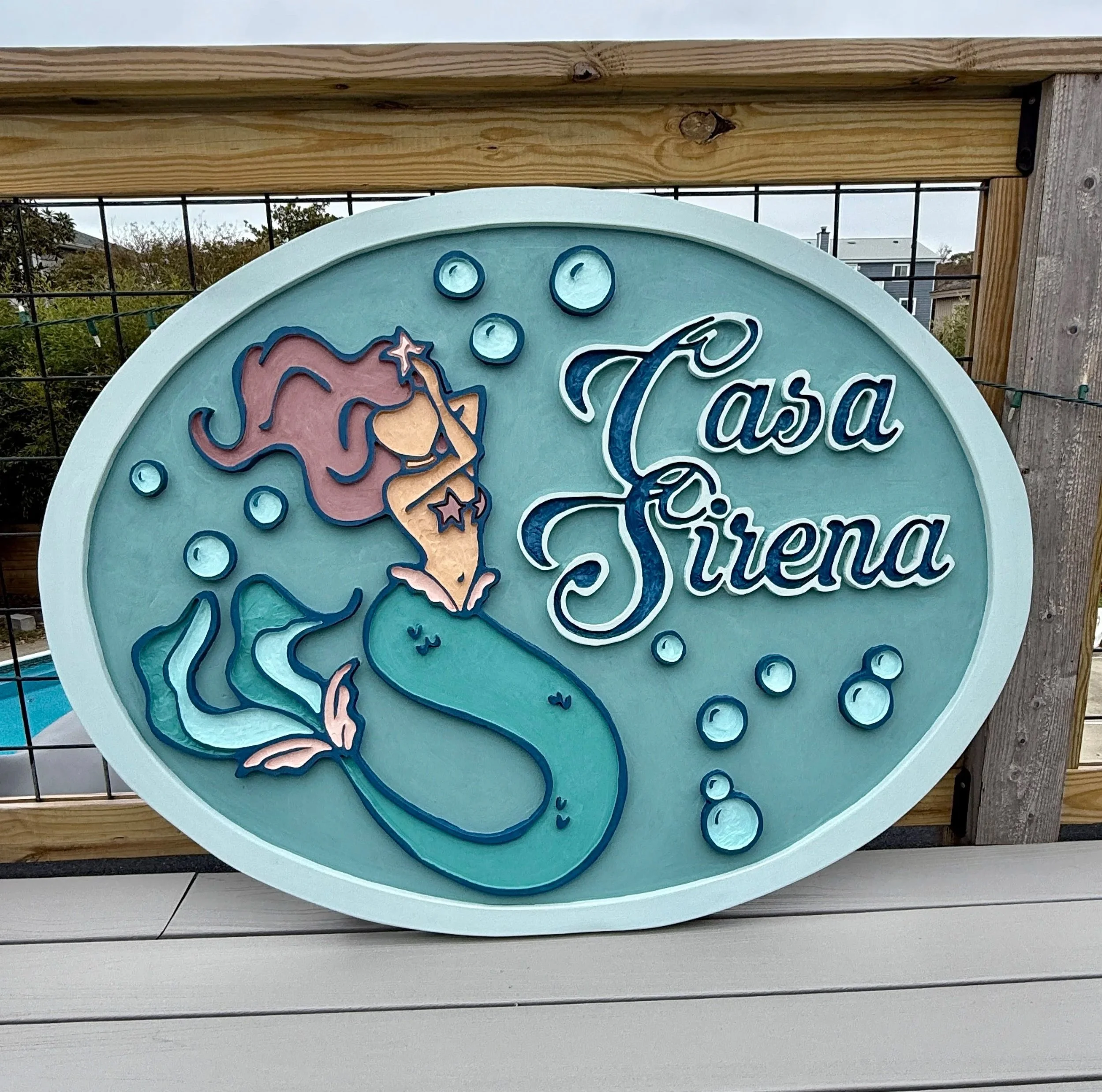 A round sign with a mermaid and text that reads 'Tasia.' The sign is mounted on a wooden frame with black metal fencing and a background of trees and buildings.