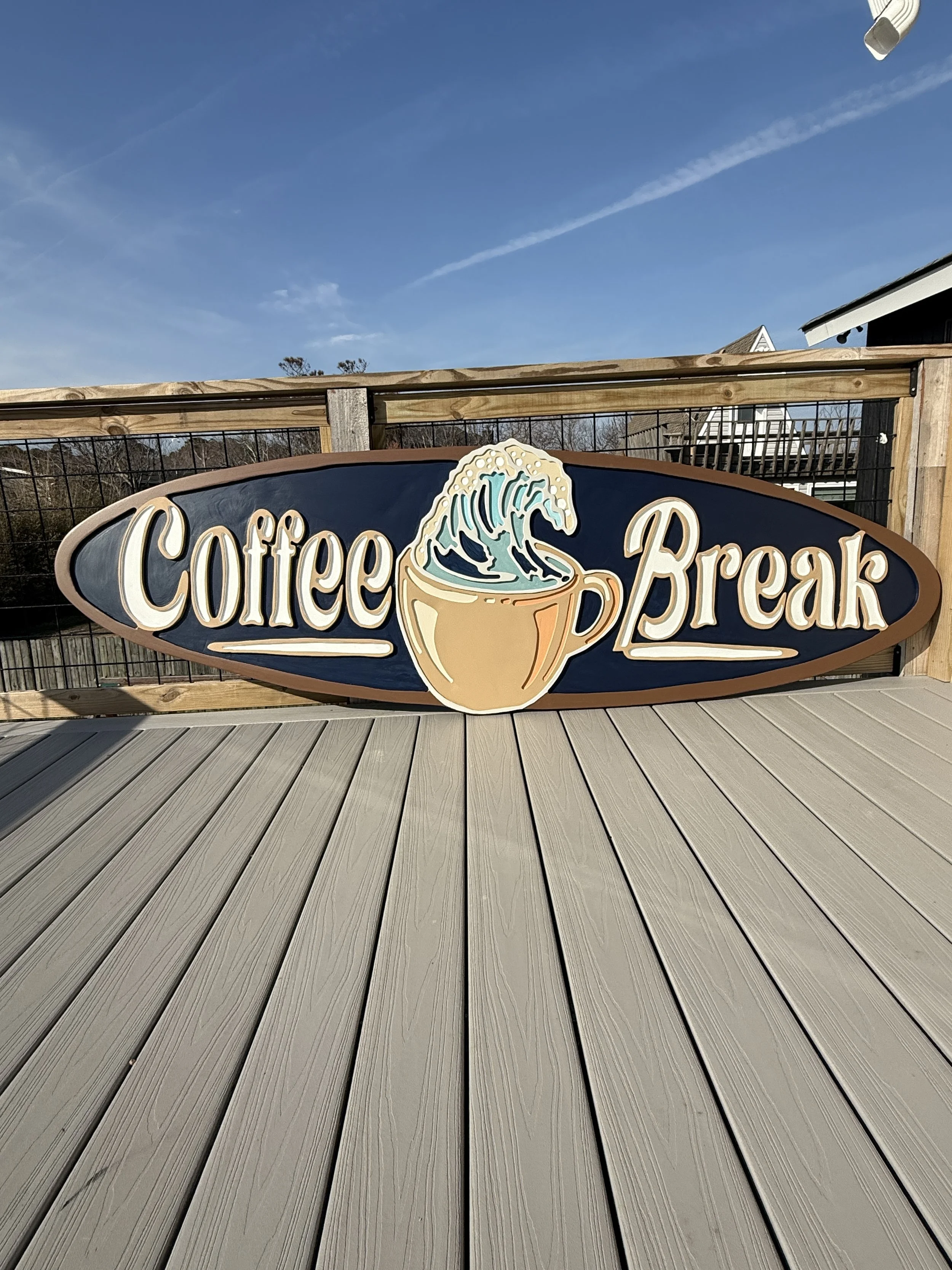 Sign for Coffee Break with a coffee cup and wave design, mounted on a wooden fence on a patio or deck, under a clear blue sky.