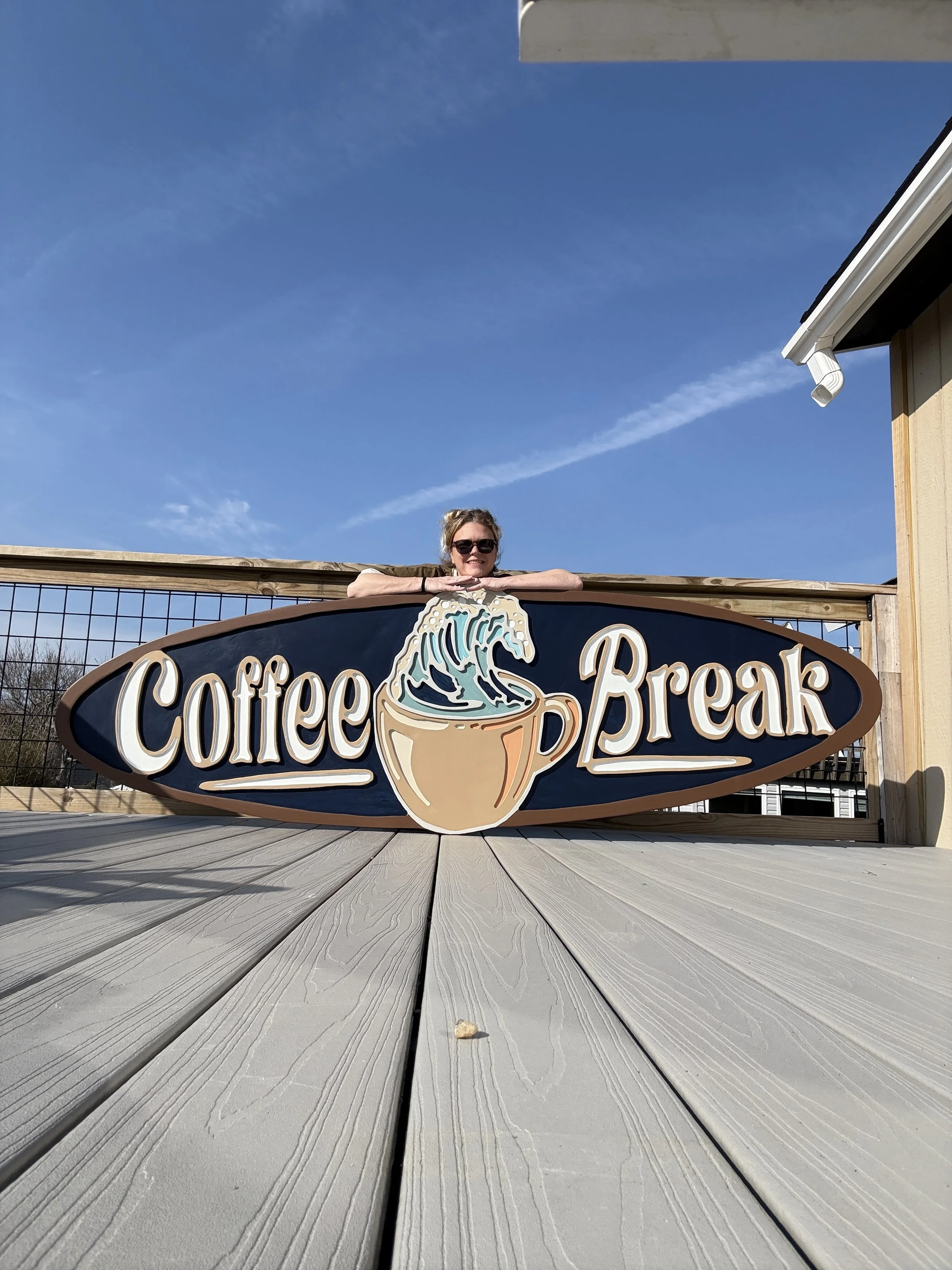 A woman wearing sunglasses leaning on a sign that reads 'Coffee & Break' with a stylized coffee cup and a wave, standing behind a wooden fence on a deck with gray planks, under a clear blue sky.