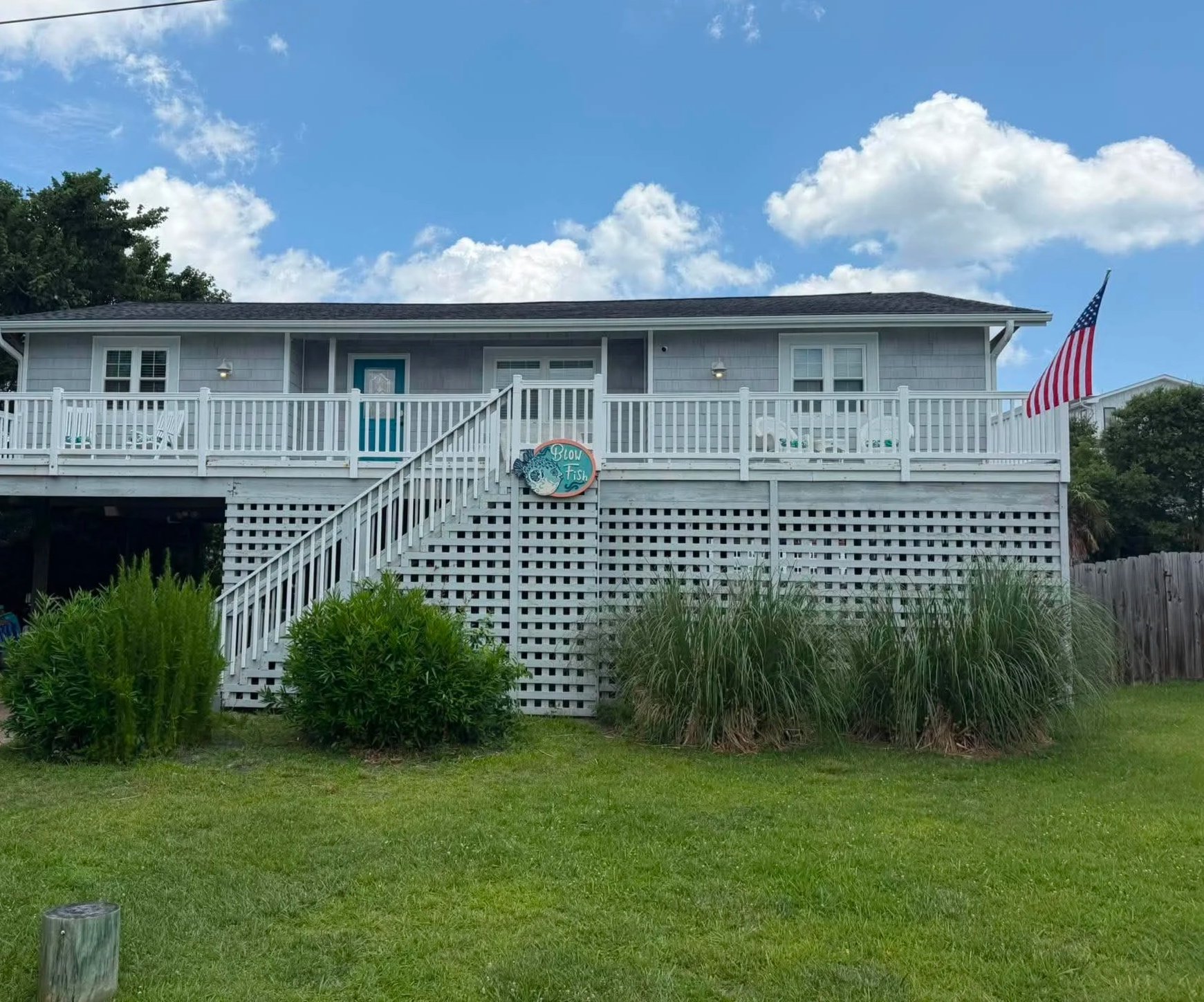A two-story house with a light blue exterior, white railing, and a staircase leading to a front porch with chairs. An American flag is displayed on the right side of the house, and a sign reading 'Blow Fish' is hanging near the staircase. The yard is green with bushes and grass, under a partly cloudy blue sky.