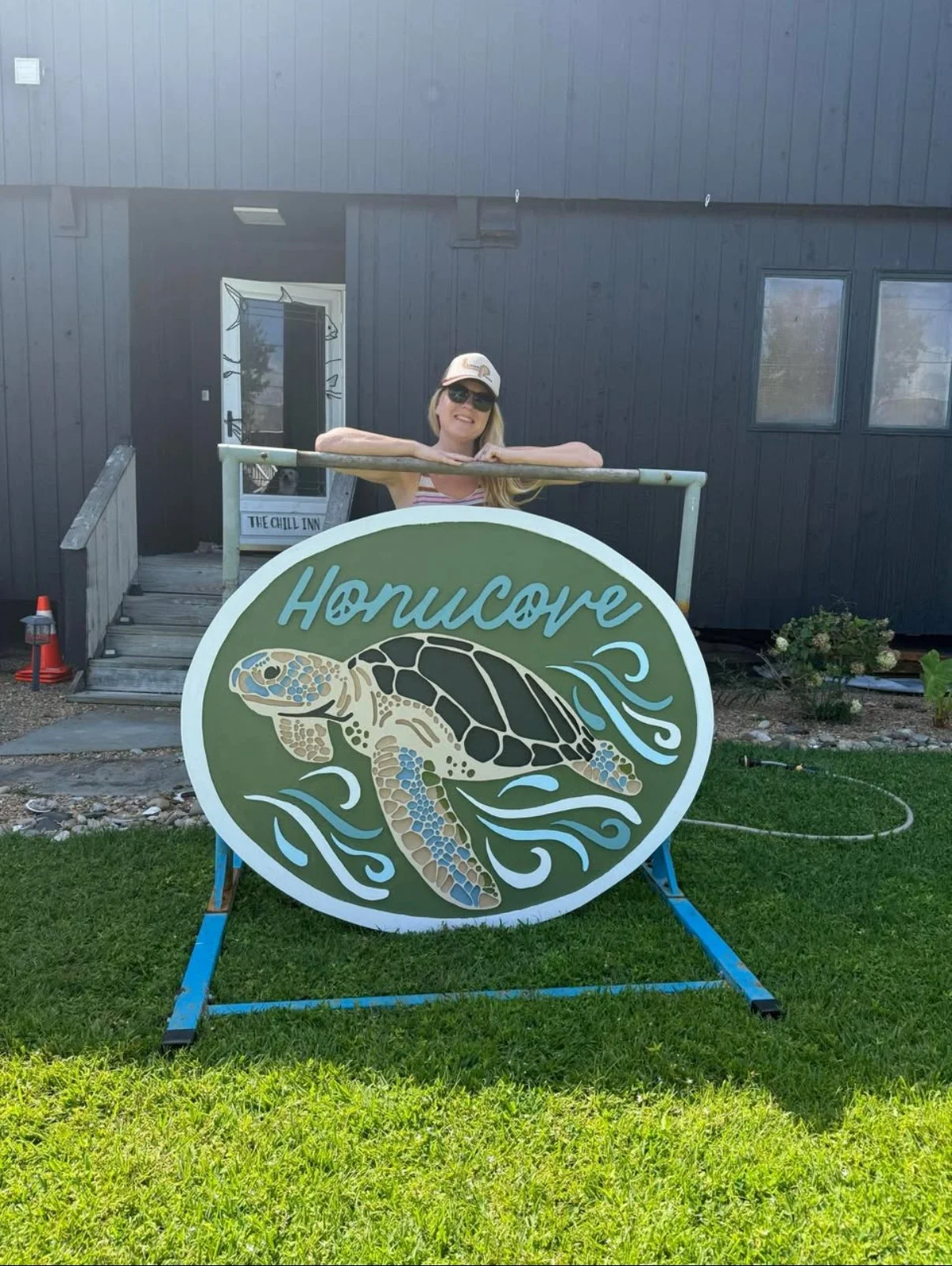 Woman standing behind a large turtle sign with the word 'Honusore' on it, in front of a dark building with a small garden and a pathway.