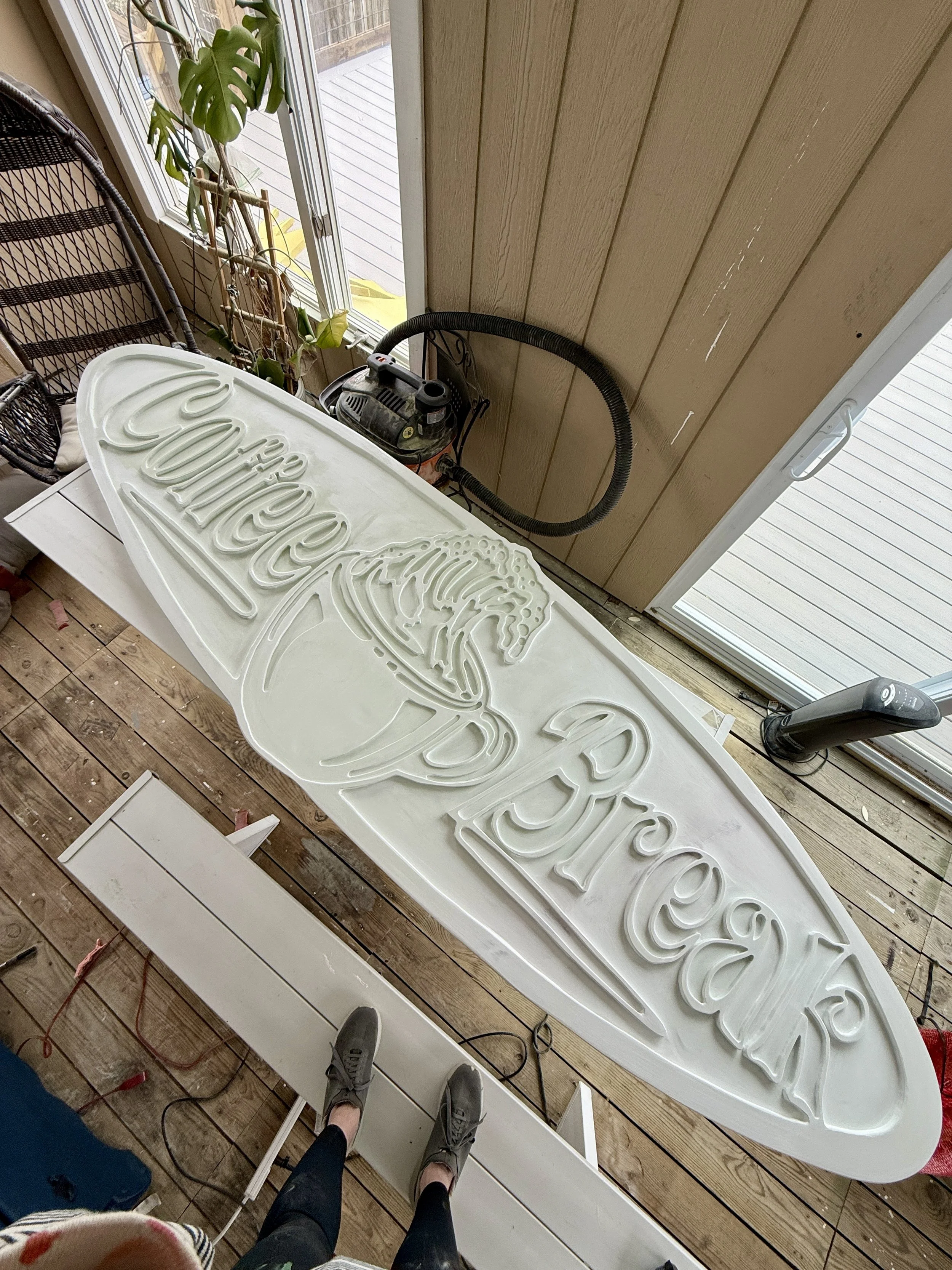 A person standing on a wooden porch looking down at a white plastic sign with raised letters that spell out "CocaCola" in cursive, while a vacuum cleaner is nearby and a potted plant sits by a sliding glass door.
