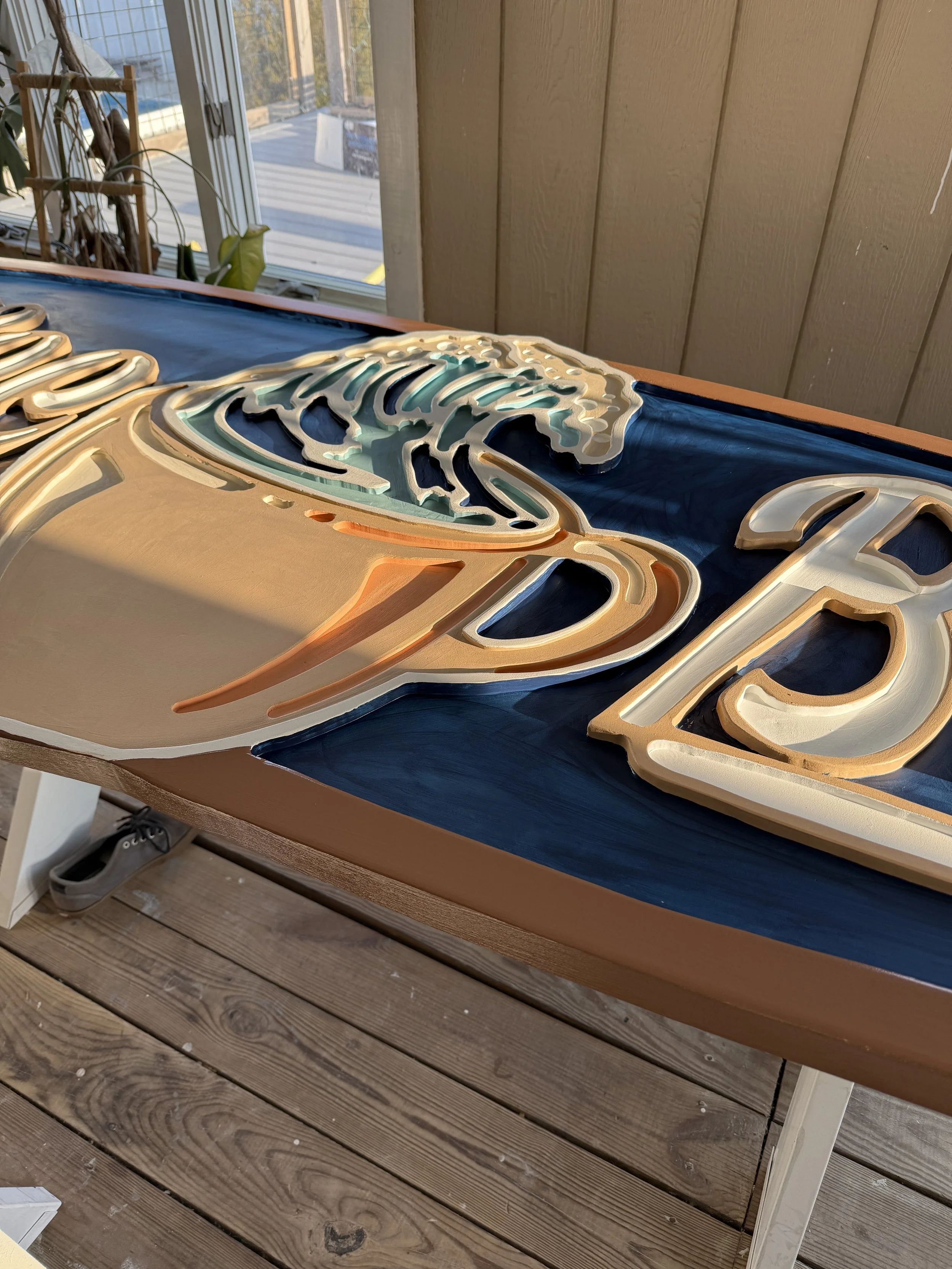 Close-up of a decorative sign with layered wooden cutouts on a table near a window with plants and a wooden wall in the background.