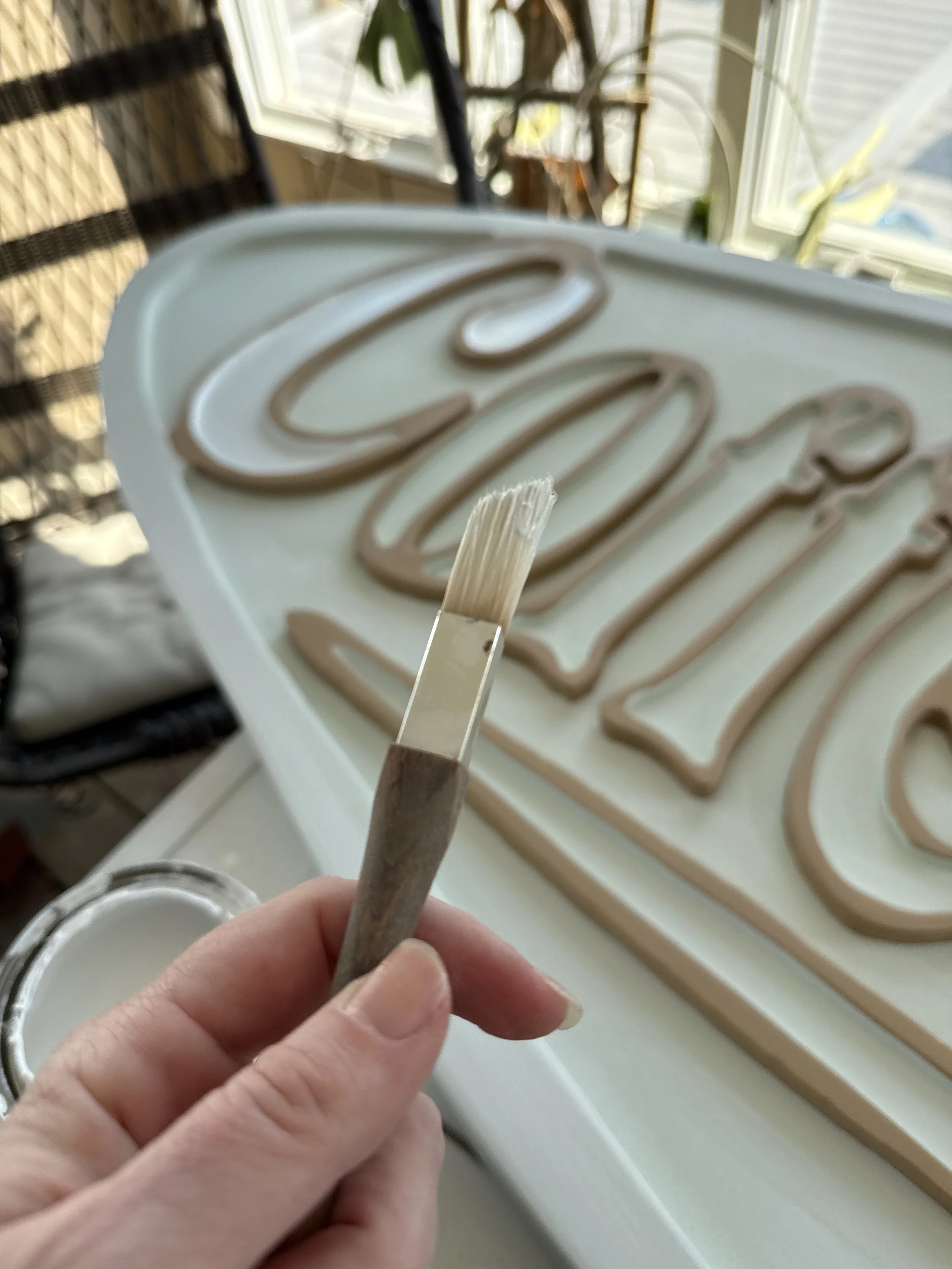 Close-up of a hand holding a small flat brush with beige or light brown icing, being used to create a decorative design on a tray of piping gel with beige or light brown icing. In the background, there is a window with blinds and some houseplants.