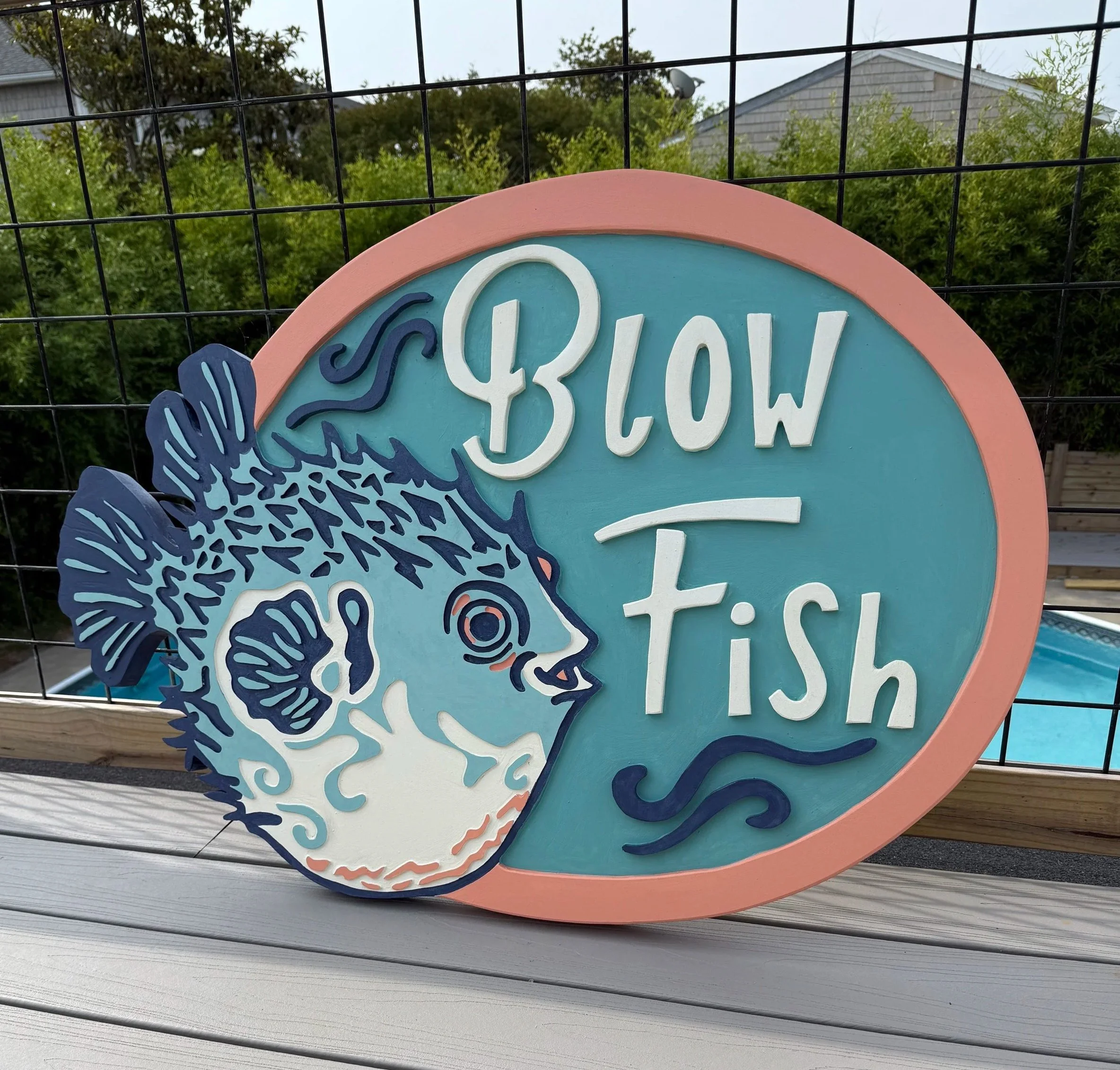 Colorful decorative sign with the word 'SHOW' in white, surrounded by floral and wave patterns, attached to a black wire fence with trees and houses in the background.