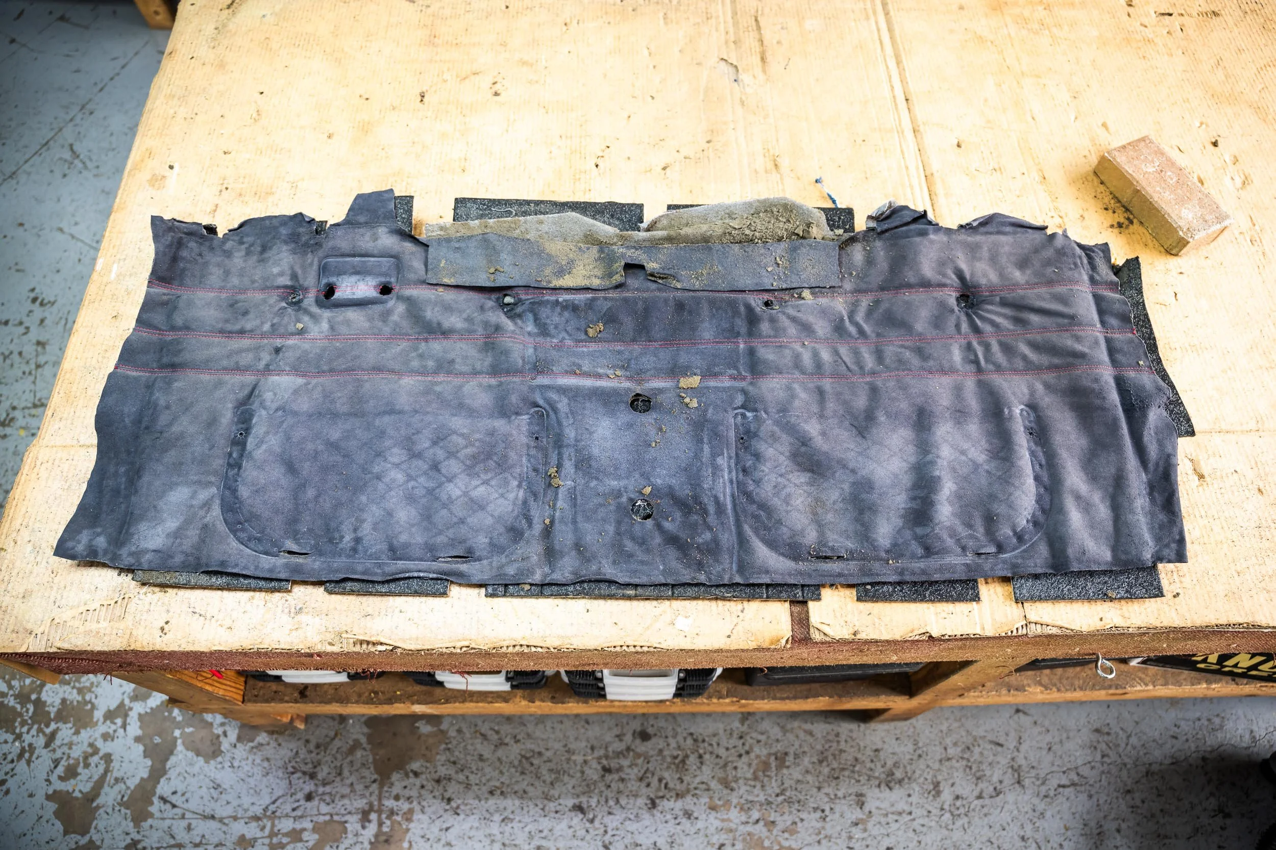 A worn and torn black rubber car trunk weatherstrip with dirt and damage, placed on a wooden workbench, next to a small brick and in a workshop environment.