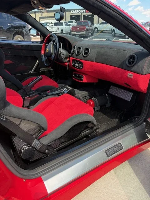 The interior of a sports car with a racing seat, red and black interior accents, and a manual gear shift, parked at a dealership lot with multiple cars in the background.