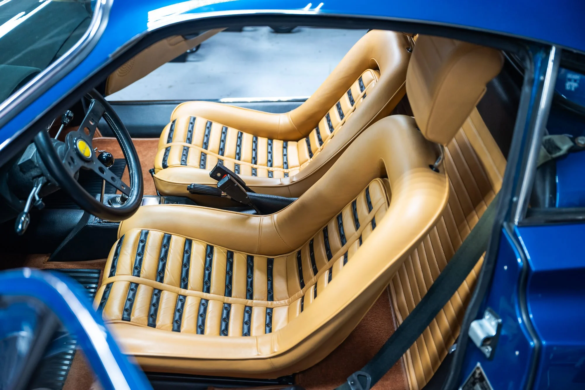 Interior of a classic blue sports car, showing tan leather seats with black straps, a black steering wheel with a yellow emblem, and brown carpet flooring.