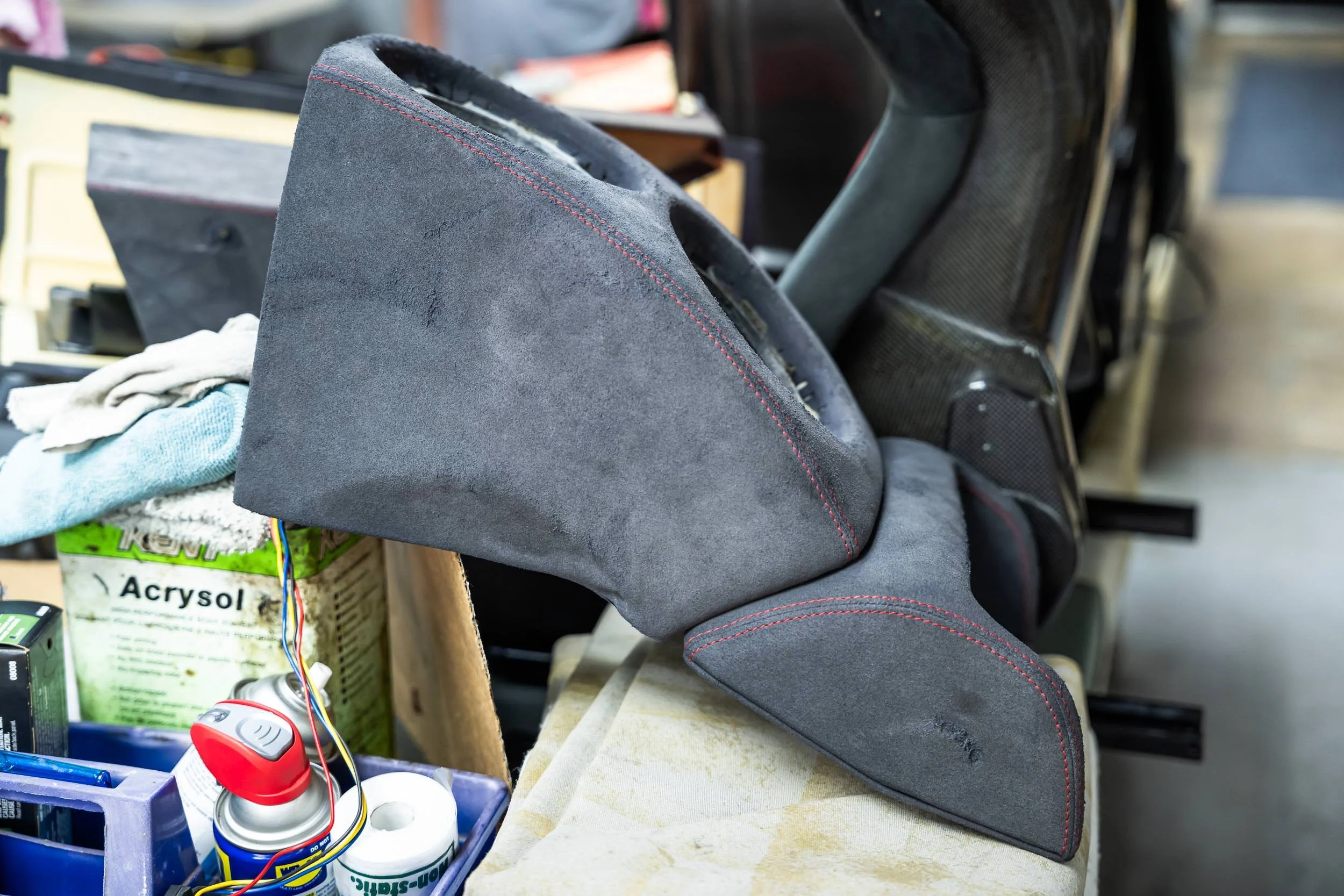 Close-up of a partially disassembled gray leather automotive seat with red stitching, surrounded by various tools, wires, and supplies in a workshop.