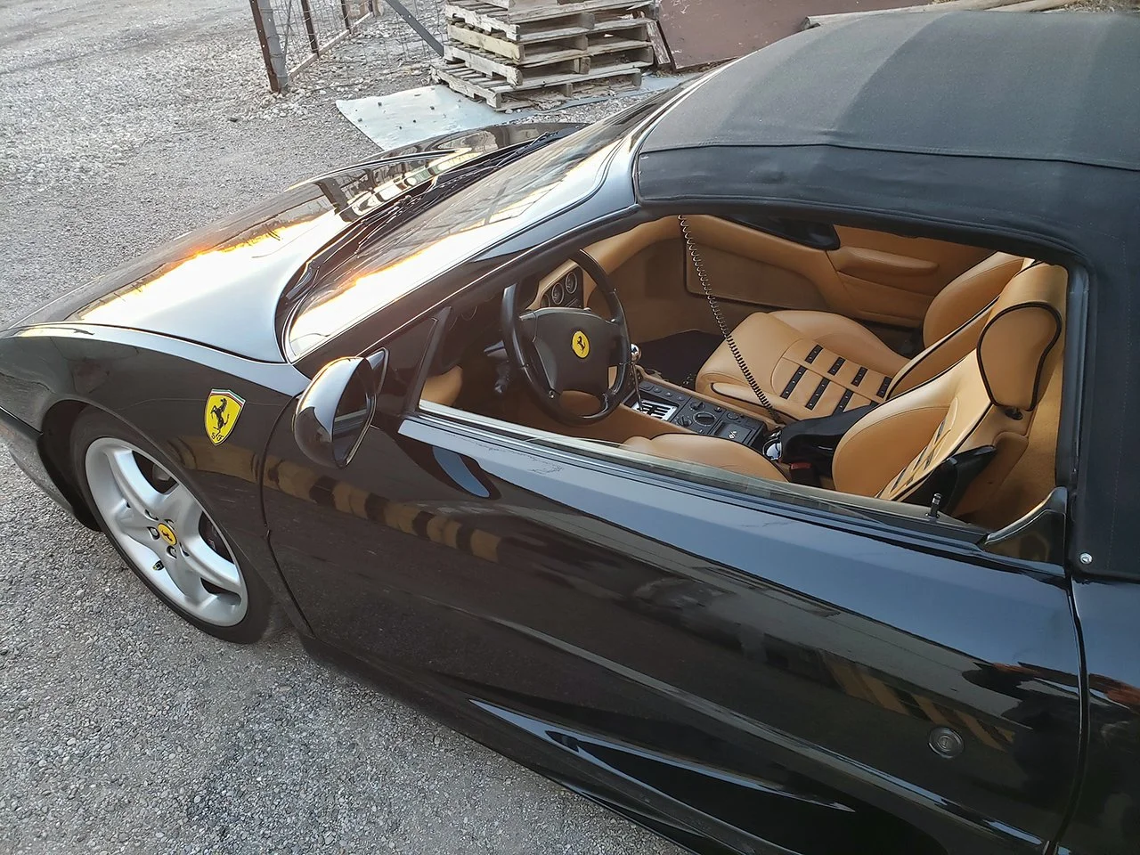Black Ferrari convertible with tan leather interior, parked on gravel, showing the interior details and the Ferrari emblem on the wheel and body.