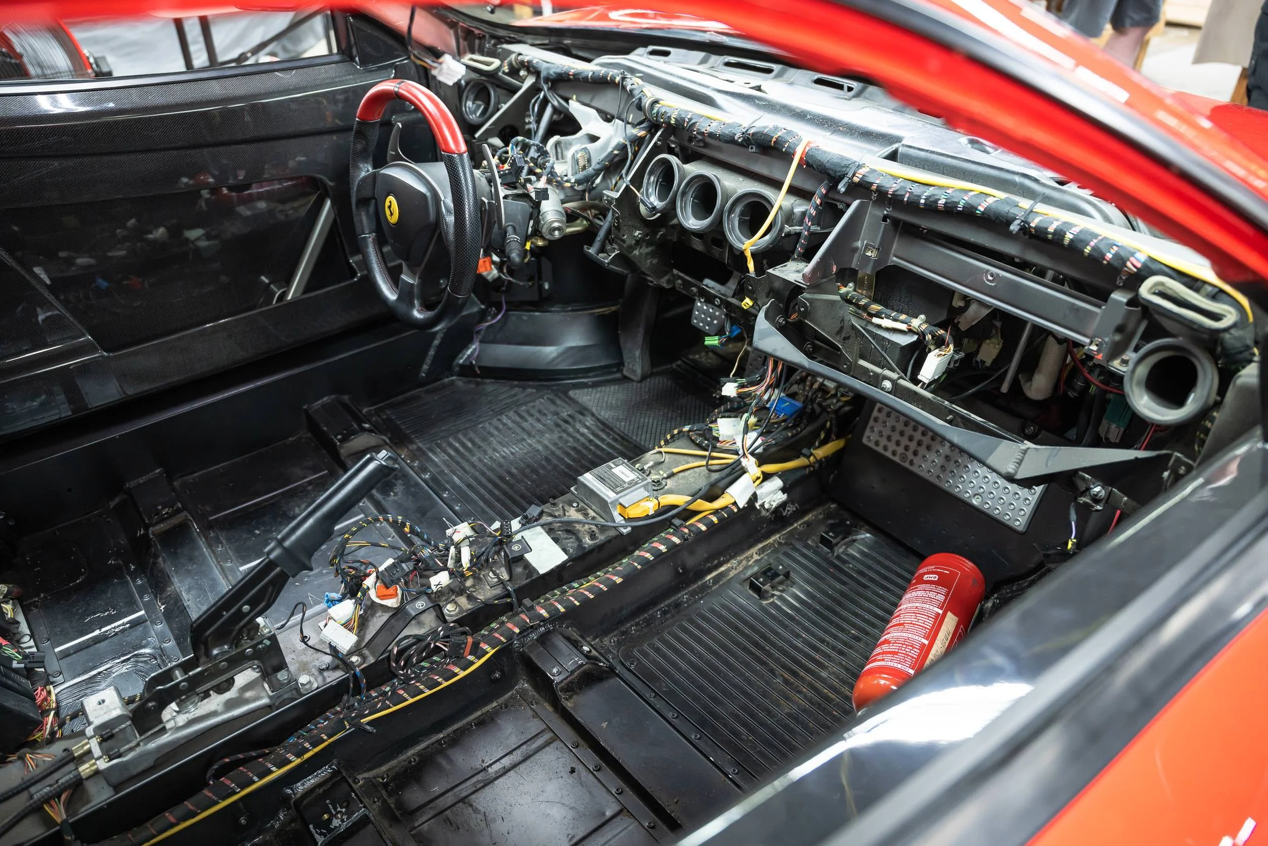 The interior of a race car with the dashboard disassembled, exposing wiring, the steering wheel with the Ferrari logo, and fire extinguisher, indicating ongoing maintenance or repair.
