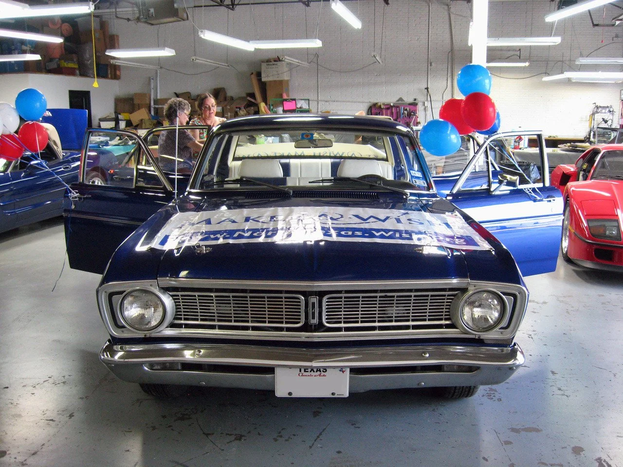 Black vintage car in an indoor garage decorated with red, white, and blue balloons, with people talking in the background.
