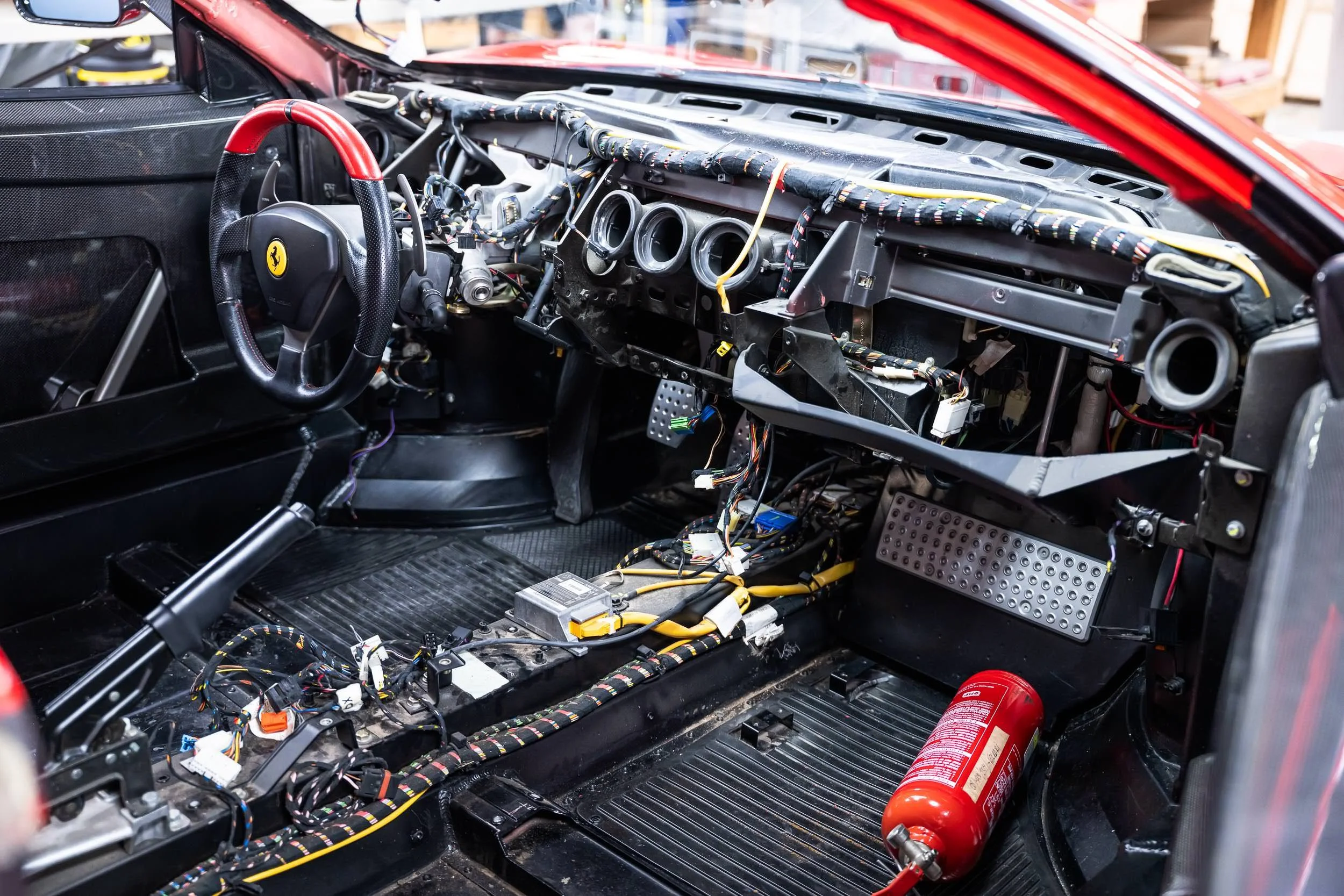 Interior of a race car under construction, showing wiring and dashboard without instruments, with a Ferrari logo on the steering wheel, and a fire extinguisher on the floor.