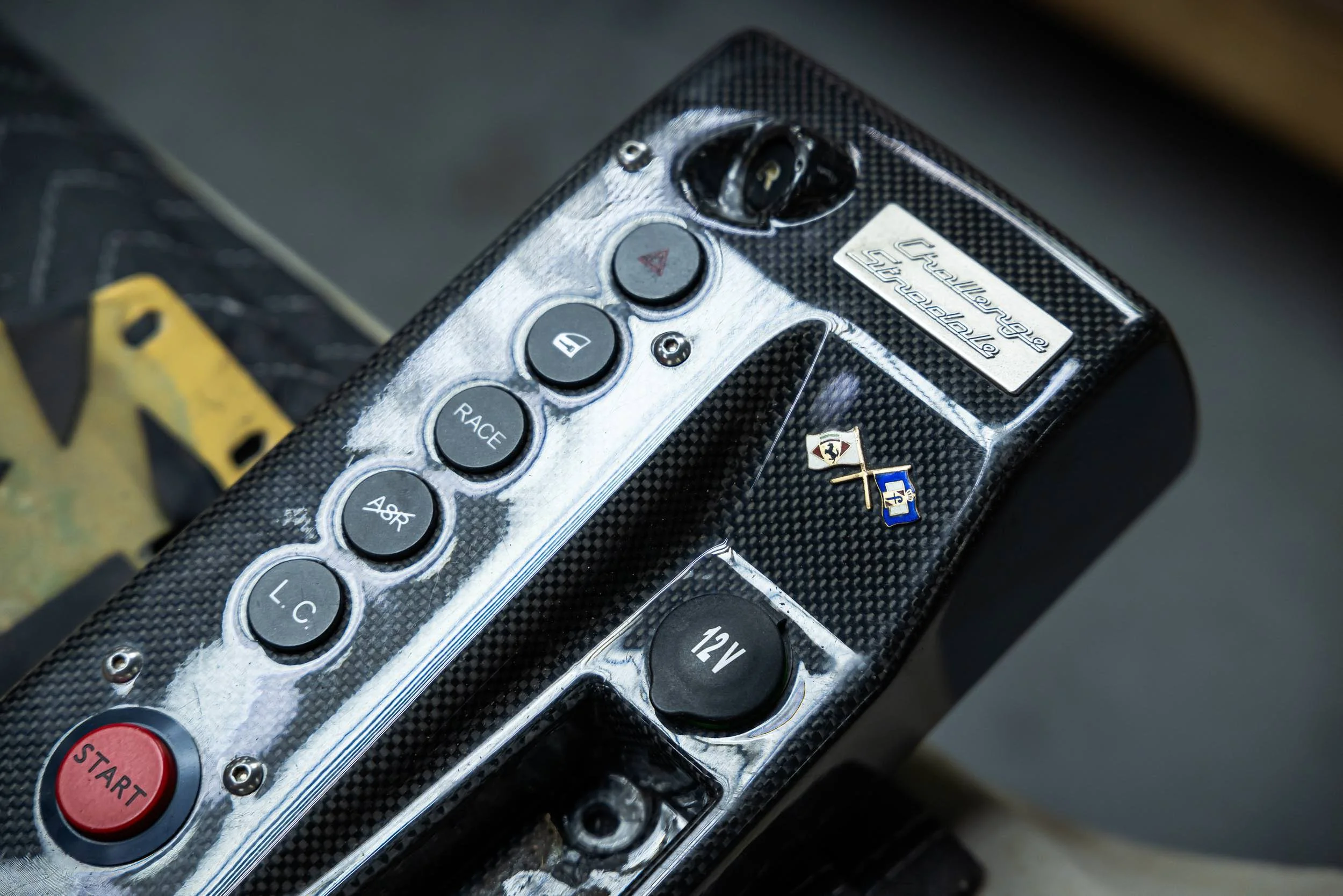 Close-up of a racing car remote control placed on a carbon fiber surface, with various buttons including 'START', 'RACE', and labeled controls, alongside a small Ferrari badge.