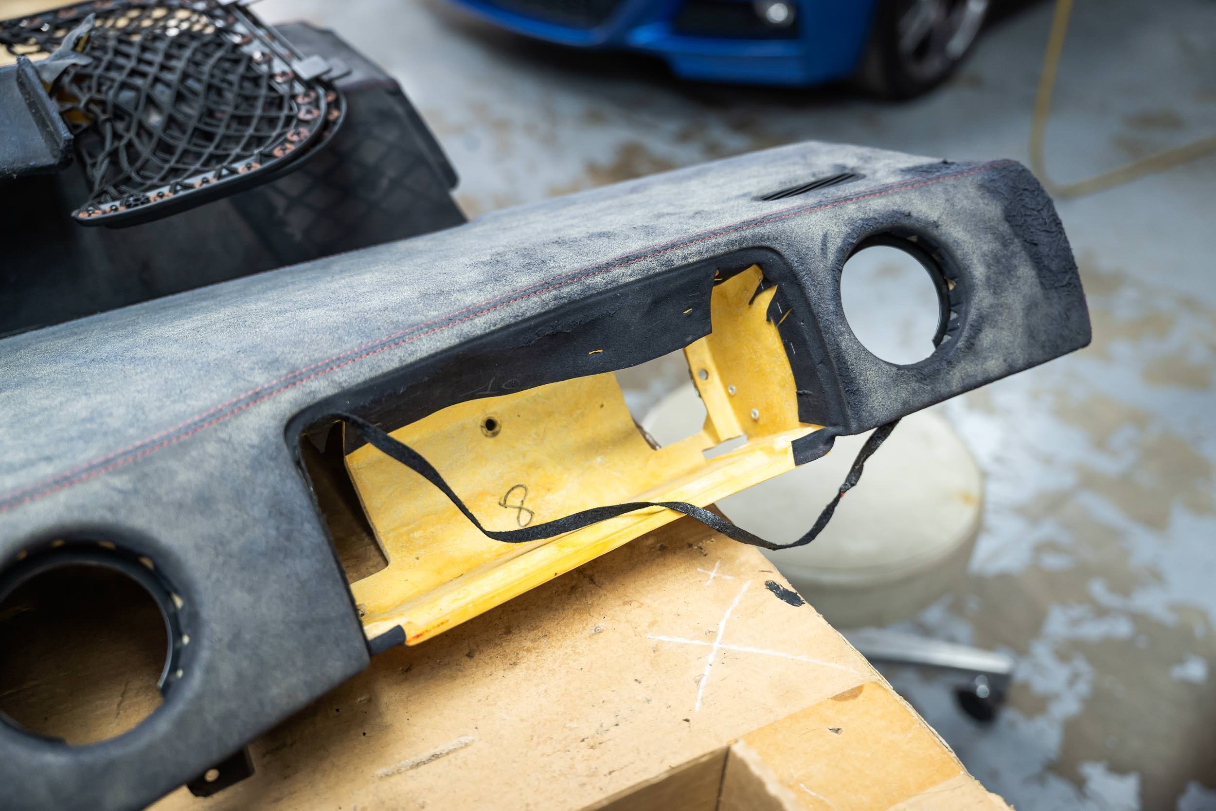 Close-up of a speaker box with a yellow speaker inside, placed on a wooden surface. The speaker box appears to be disassembled or under repair in a garage or workshop setting.