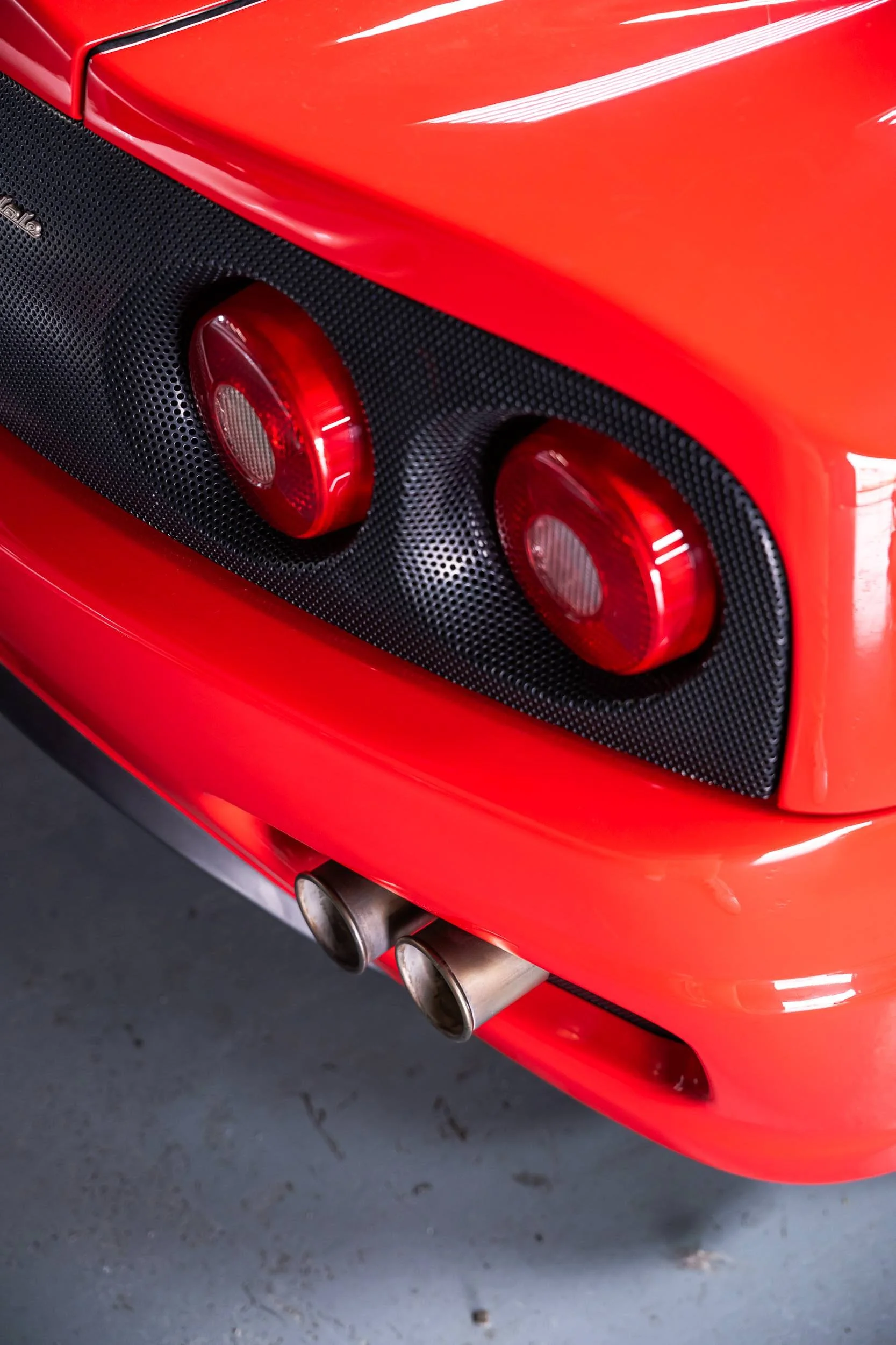 Close-up of the rear section of a red sports car, showing two circular red tail lights, a carbon fiber panel, and dual exhaust pipes.