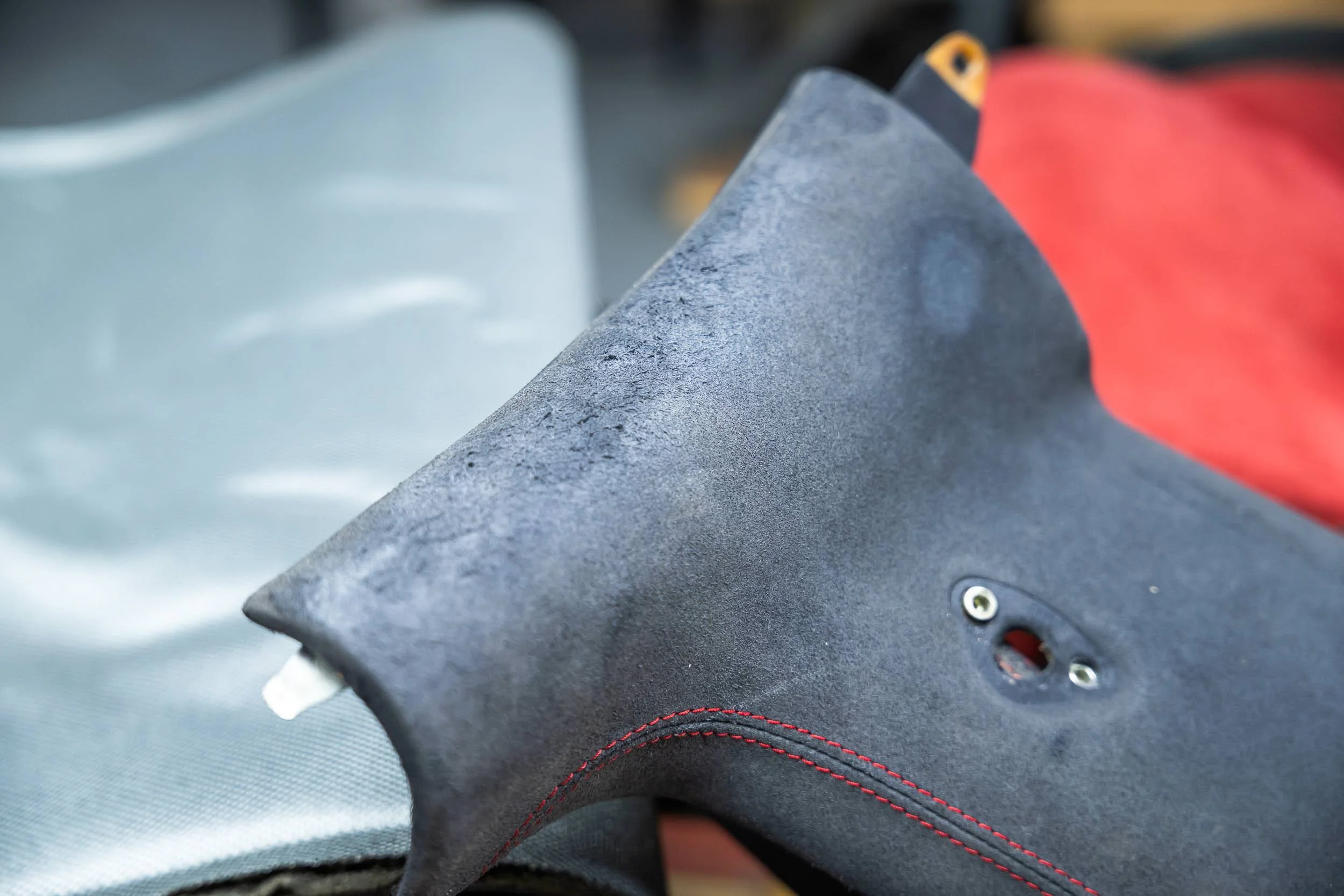 Close-up of a motorcycle seat with black material, visible red stitching, and a small part of a red backrest or cover, with a metallic component nearby.