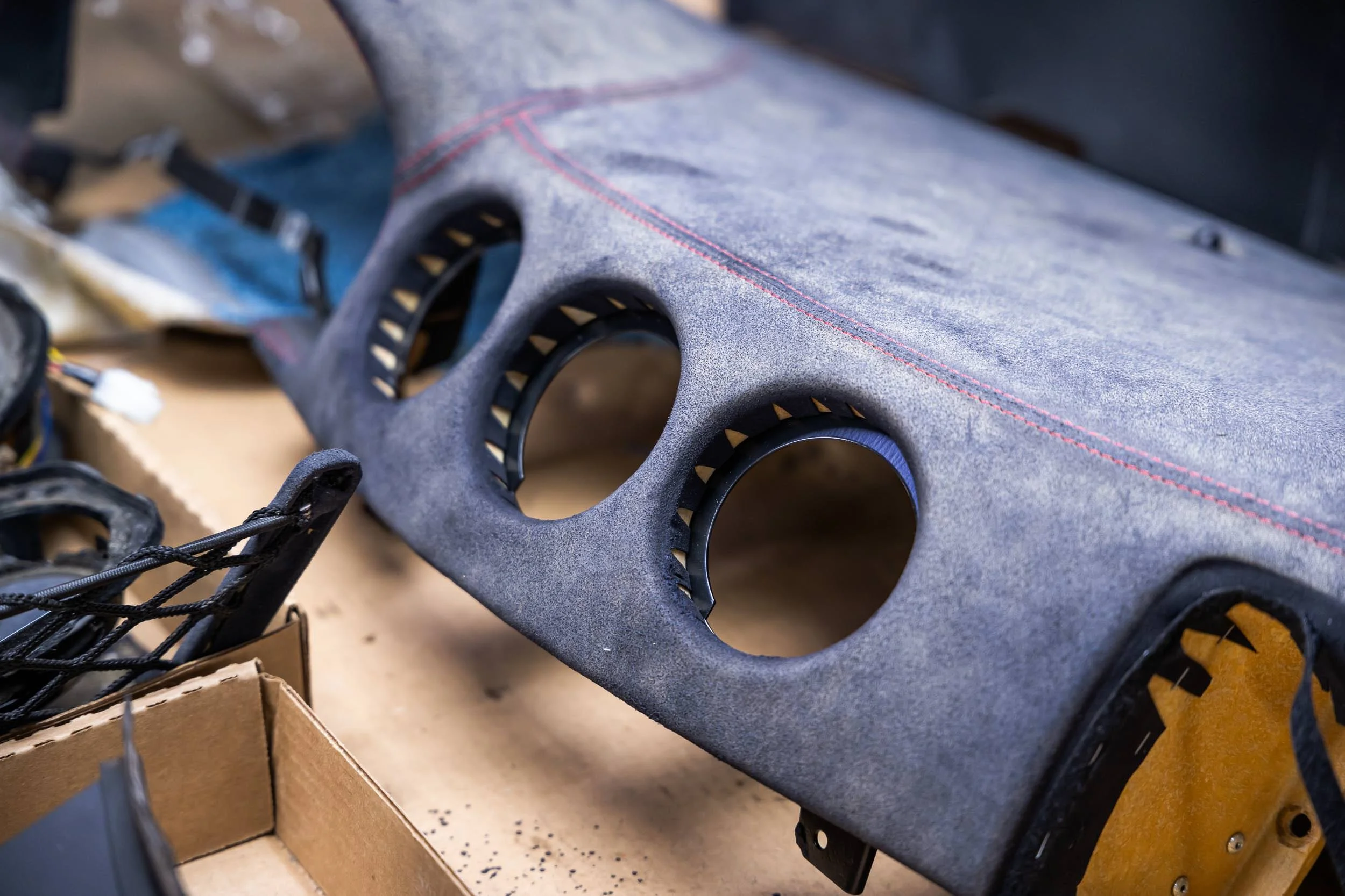 Close-up of a gray car dashboard with circular air vents and red stitching detail, surrounded by various car parts and tools, on a cluttered workbench.