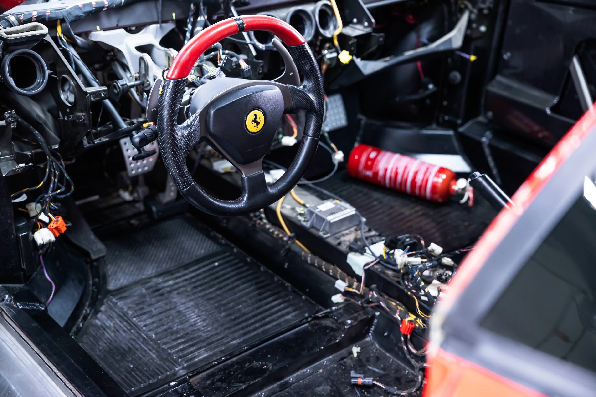 Inside of a race car with the steering wheel attached, showing an incomplete dashboard and exposed wiring, circuitry, and fire extinguisher.