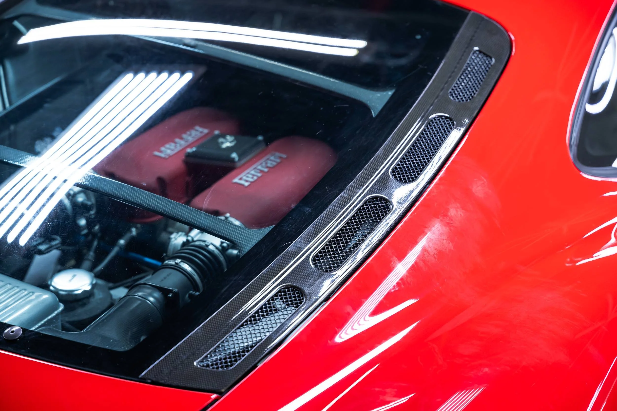 Close-up of a red sports car engine viewed through a glass window on the hood, with visible Ferrari branding on the engine components.