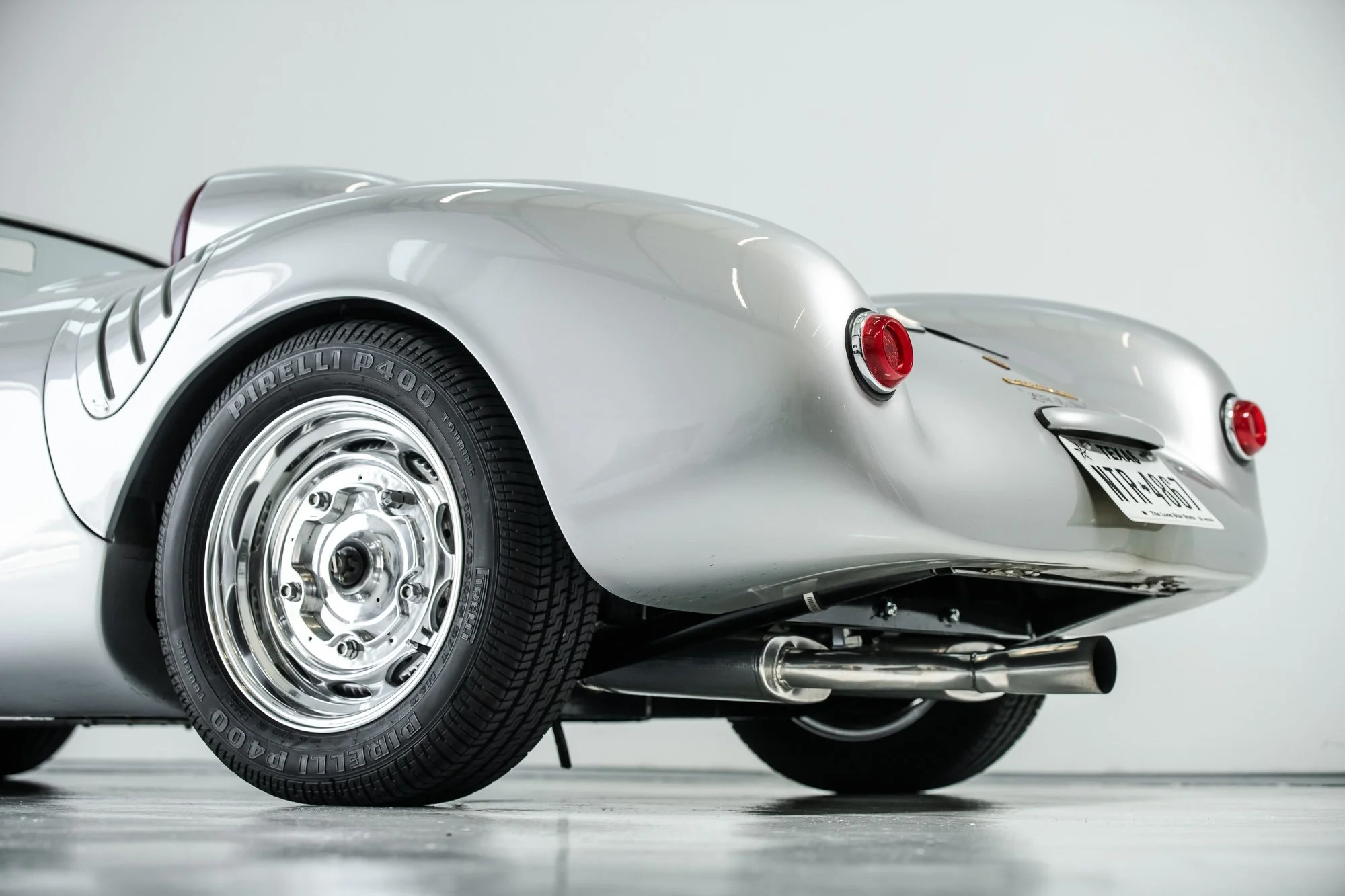 Close-up of the rear of a silver vintage sports car, showing a Pirelli P400 tire, classic round taillights, and a visible exhaust pipe on a plain background.